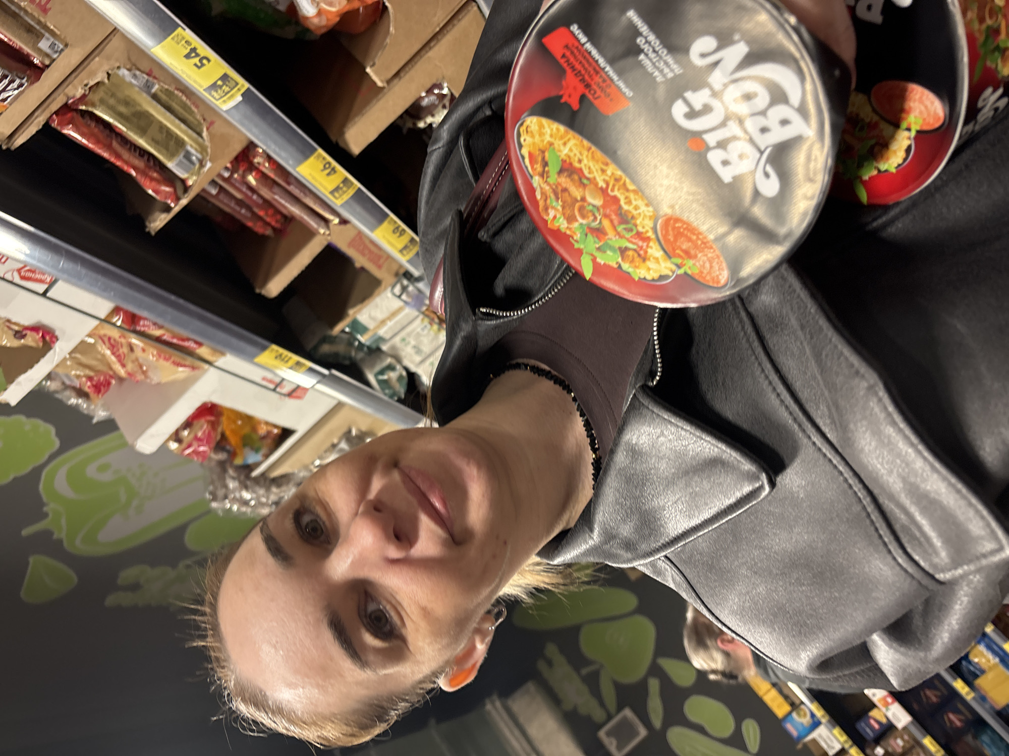 A woman is captured in a selfie-style photograph inside what appears to be a supermarket or grocery store. She has light-colored hair and is wearing a black leather jacket over a dark-colored top. She holds two cups of "BIG BON" instant noodles, one in front of the other, showcasing their packaging. Both noodle cups feature an image of prepared noodles with beef, vegetables, and sauce. The woman looks directly at the camera with a neutral expression, possibly a subtle smile.
The background reveals typical supermarket shelving, stocked with various packaged goods. Yellow price tags are visible on the shelves, displaying prices such as "54.99," "46.99," and "69.99," likely in Rubles given the location context. A dark wall or floor area behind the woman is adorned with light green, stylized outlines of vegetables, including peppers and beans. Part of another person's head is faintly visible in the lower right background.
Visible text on the noodle cups includes the brand "BIG BON," and in Cyrillic: "ЛАПША БЫСТРОГО ПРИГОТОВЛЕНИЯ" (Instant Noodles), "ГОВЯДИНА" (Beef), and "ОРИГИНАЛЬНЫЙ ВКУС" (Original Taste). The presence of Cyrillic text on both the product packaging and price tags, along with the "BIG BON" brand common in the region, supports the provided location context of Russia. The exact time of day and external weather conditions are not discernible from the indoor setting.