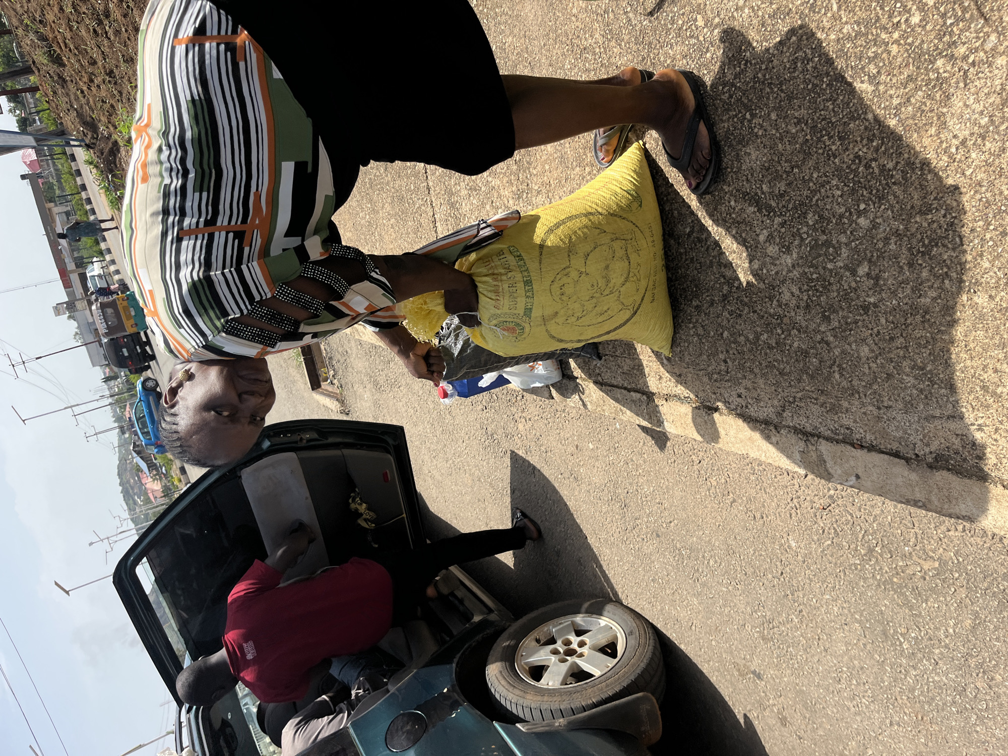 In Bukuru, Nigeria, a woman in a colorful striped shirt and black skirt is bending over to pick up a large yellow bag labeled "SUPER STARTER." She is wearing dark flip-flops. Beside her is a dark green SUV with its back door open, and a man in a red shirt is standing inside the vehicle. The ground is paved with asphalt, and a concrete curb runs alongside it. In the background, there are streetlights, other vehicles, and buildings, suggesting an urban or semi-urban environment. The scene appears to be taking place outdoors during the daytime, with strong sunlight casting shadows.