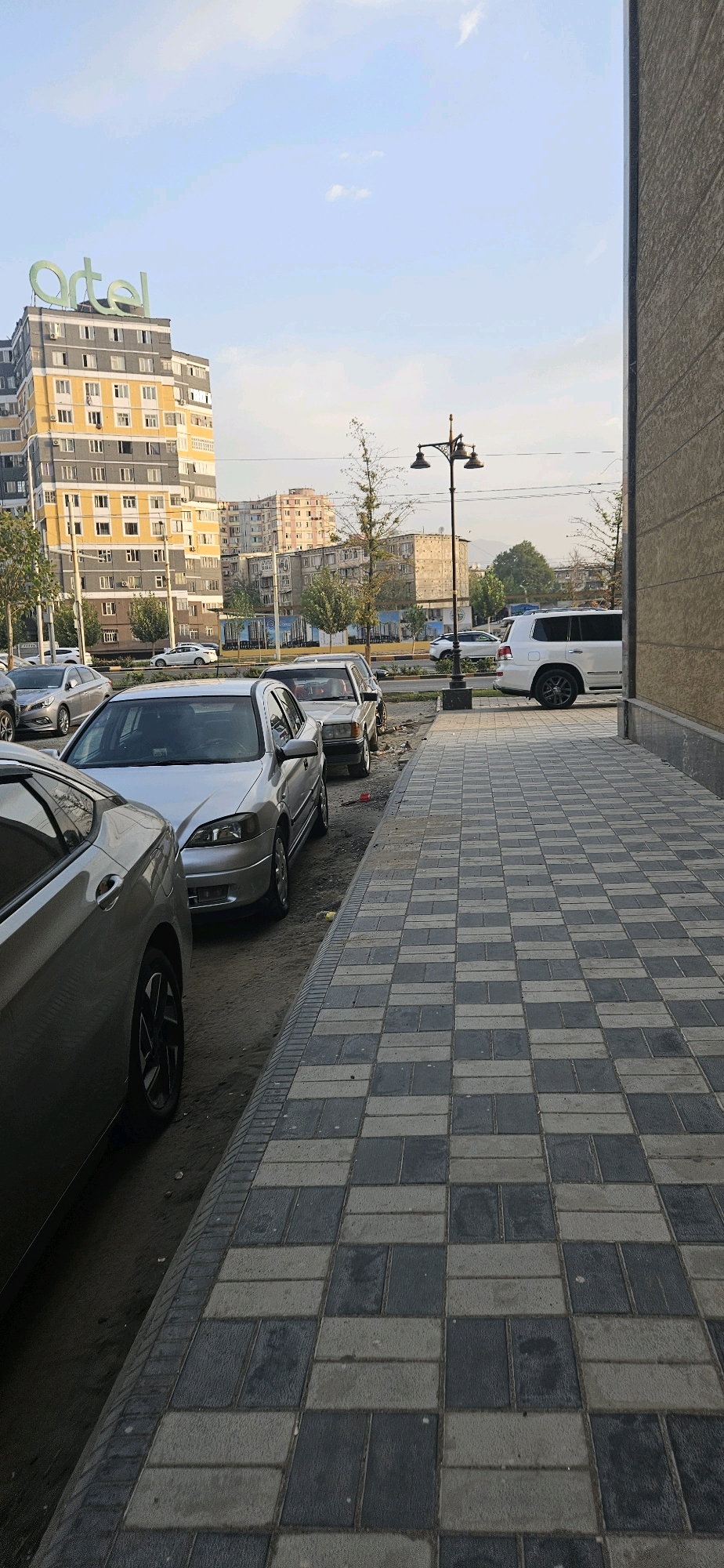 The image depicts a street scene in Dushanbe, Tajikistan, likely in a commercial or residential area. In the background, a tall building with the green "ARTEL" logo prominently displayed on its rooftop stands out. The building has a yellow and grey facade with numerous windows. Several other residential buildings of varying heights and architectural styles are visible further in the background, suggesting an urban environment.
In the foreground and mid-ground, multiple cars are parked along the side of the street. These include a silver sedan, a white SUV, and other various vehicles. A street lamp with two light fixtures stands near the sidewalk.
The sidewalk itself is paved with rectangular interlocking tiles in a checkered pattern of grey and off-white. The area bordering the street and sidewalk appears to be a dirt or gravel shoulder with some scattered debris. The sky is visible in the upper portion of the image and appears to be partly cloudy. The overall impression is a typical urban streetscape with parked vehicles and buildings under a bright sky.