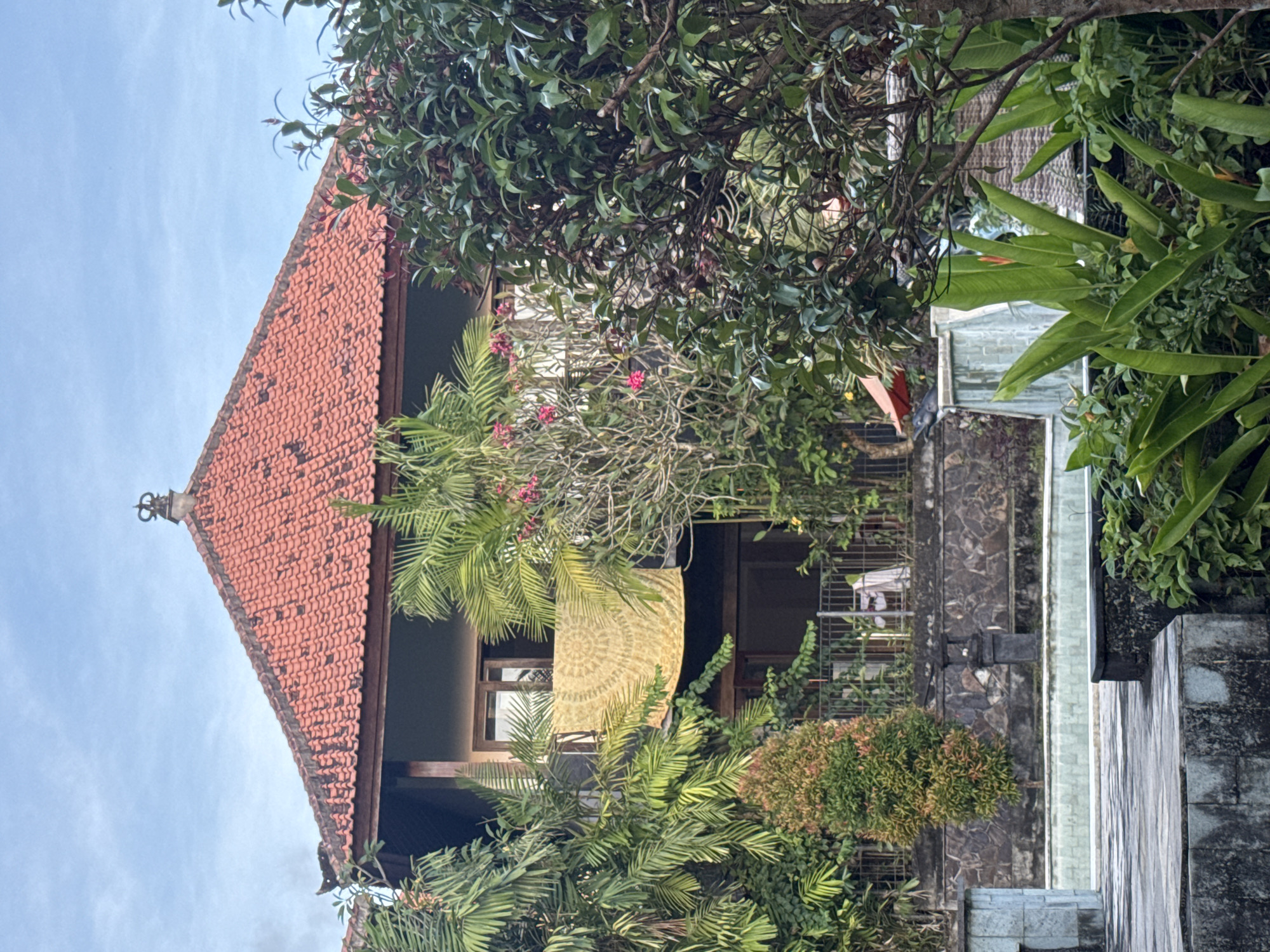 The image captures a building in Kuta Selatan, Indonesia, surrounded by lush tropical vegetation. The building features a prominent red-tiled roof with a decorative finial at its apex. Its facade is partially obscured by trees, ferns, and other greenery, including a tree with pink flowers. A patterned yellow fabric or screen hangs in front of a window. The building appears to be constructed with stone or brick elements. In the foreground, a stone wall and steps lead up to the building. The sky is visible in the upper left corner.