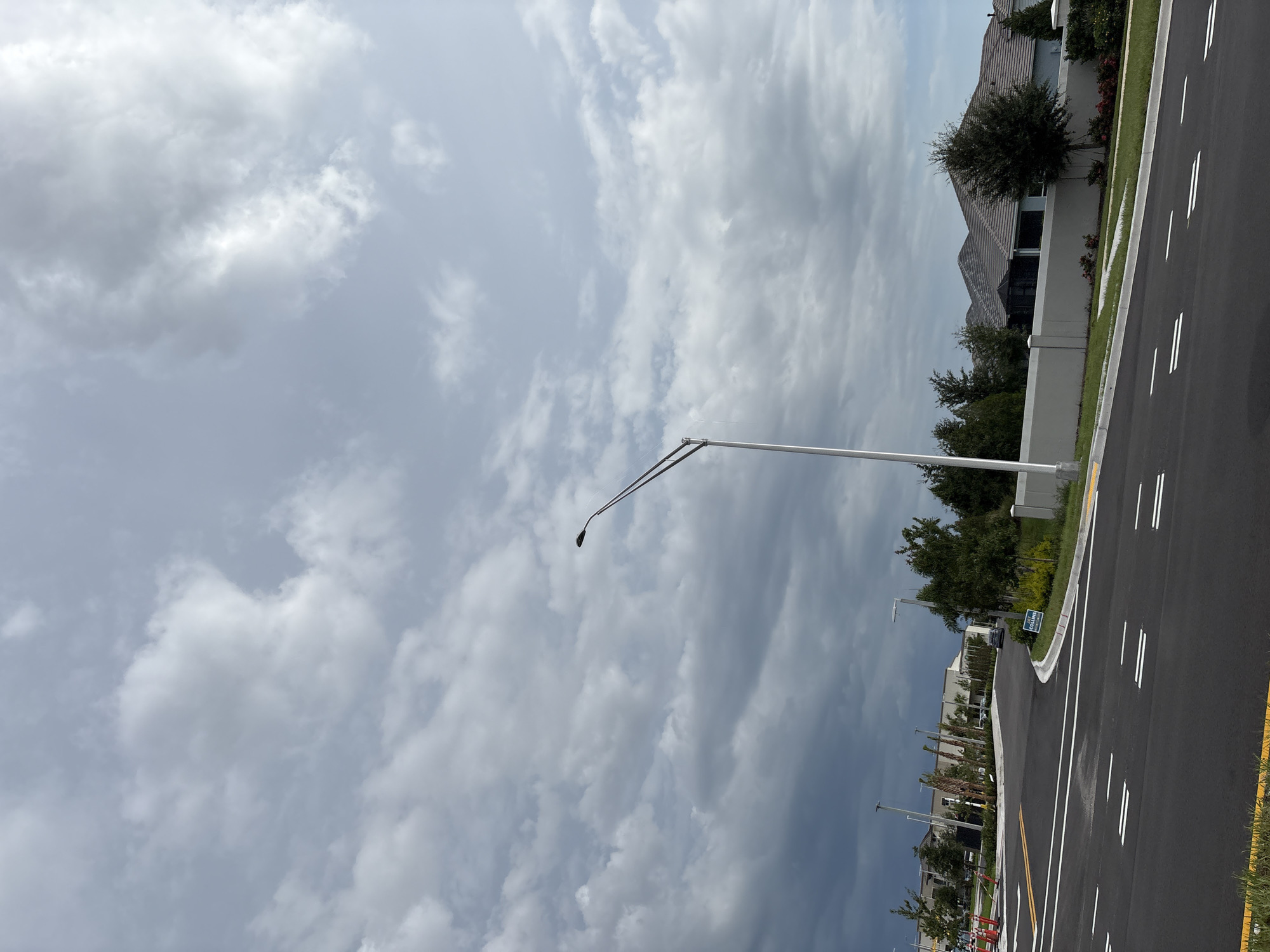 The image depicts a street scene in Bradenton, Florida, United States. A modern street lamp stands prominently in the foreground, to the right of the frame. The road is newly paved with clear white lane markings. In the background, a row of houses with tiled roofs is visible behind a low, light-colored wall. Green trees and landscaping are present, and the sky is filled with a mixture of light and dark clouds, suggesting a cloudy or potentially stormy day. A sign is partially visible on the right side of the road, with text that appears to read "COLLINS". Further in the distance, buildings and more trees line the street. The overall scene suggests a residential or developing area.