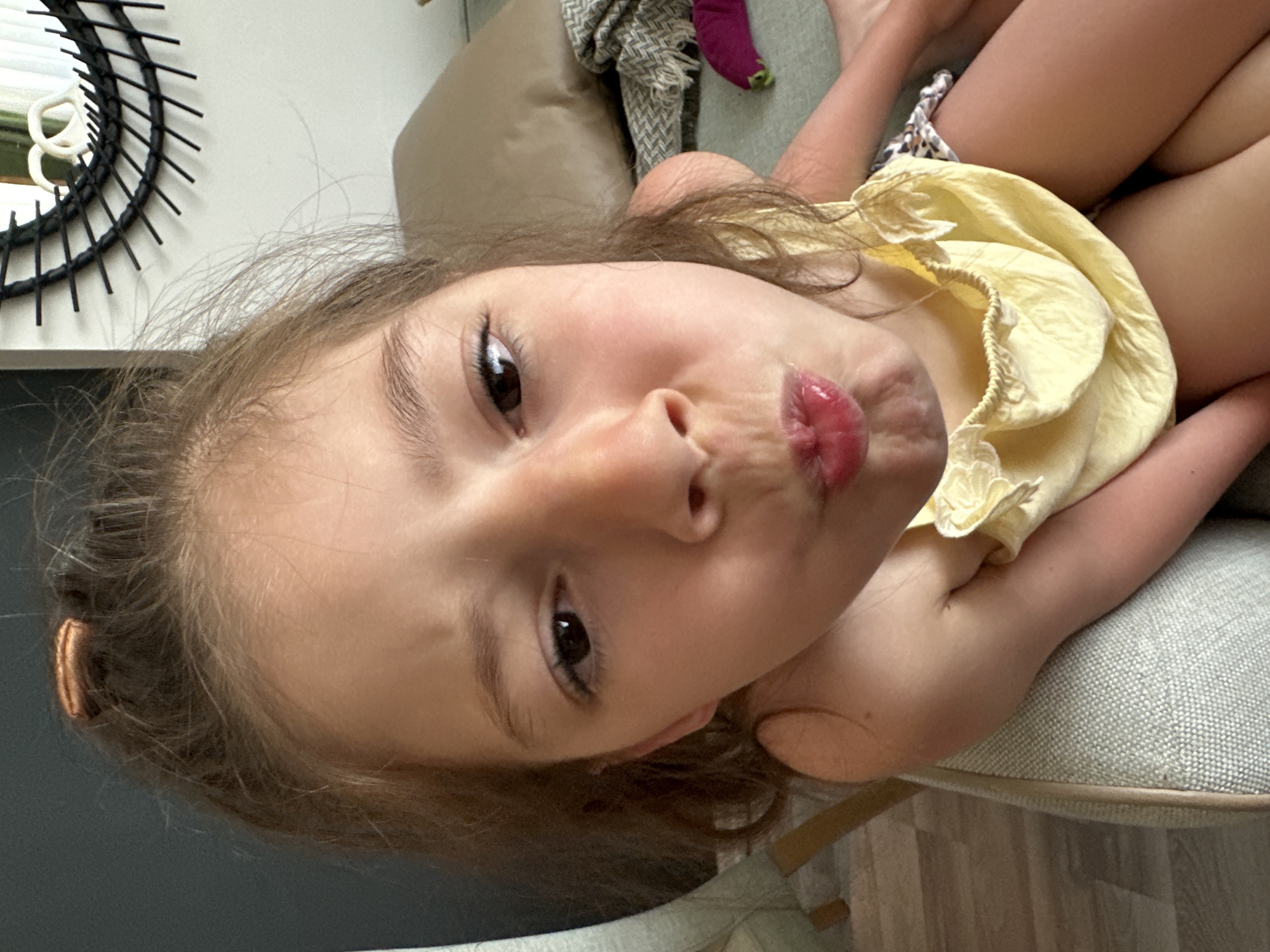 A young girl with light brown hair and brown eyes is lying on a gray cushioned chair. She has a light complexion and is wearing a yellow dress with a ruffled neckline. Her hair is pulled back with a brown clip or band. The girl is making a pouting expression with her lips. A black wicker-style mirror is visible on the wall behind her, along with a beige upholstered item, possibly a headboard or a pillow. The flooring appears to be wooden planks. There is no definitive information to confirm this photo was taken in Fethiye, Turkey.