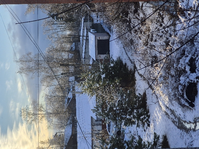The image captures a static, elevated view of a snow-covered residential or semi-rural area in Kirzhach, Russia, under a partially cloudy winter sky. The foreground exhibits a snow-covered ground interspersed with exposed earth and a winding, partially melted or muddy path extending diagonally from the lower-left. Sparse, bare tree branches occupy the extreme lower-right. Several small, snow-dusted evergreen trees are present in the foreground and midground. In the midground, a light-colored brick garage with a dark, possibly corrugated metal, roll-up door is visible, its roof covered in snow. Behind and to its left, several other single-story structures, consistent with garages or utility buildings, feature snow-covered roofs. A light-colored vehicle, likely a sedan or small SUV, is partially visible parked on a snow-covered surface near a larger building with a white, corrugated roof also bearing a layer of snow. The background reveals additional residential houses with snow-covered roofs, situated amidst numerous bare deciduous trees, which are also lightly dusted with snow. Overhead utility poles and multiple power lines crisscross the upper midground and background. The sky transitions from a bright, hazy yellowish-white on the left, suggesting a low sun angle, to a clearer blue with scattered white clouds on the right. No individuals or dynamic actions are visible.