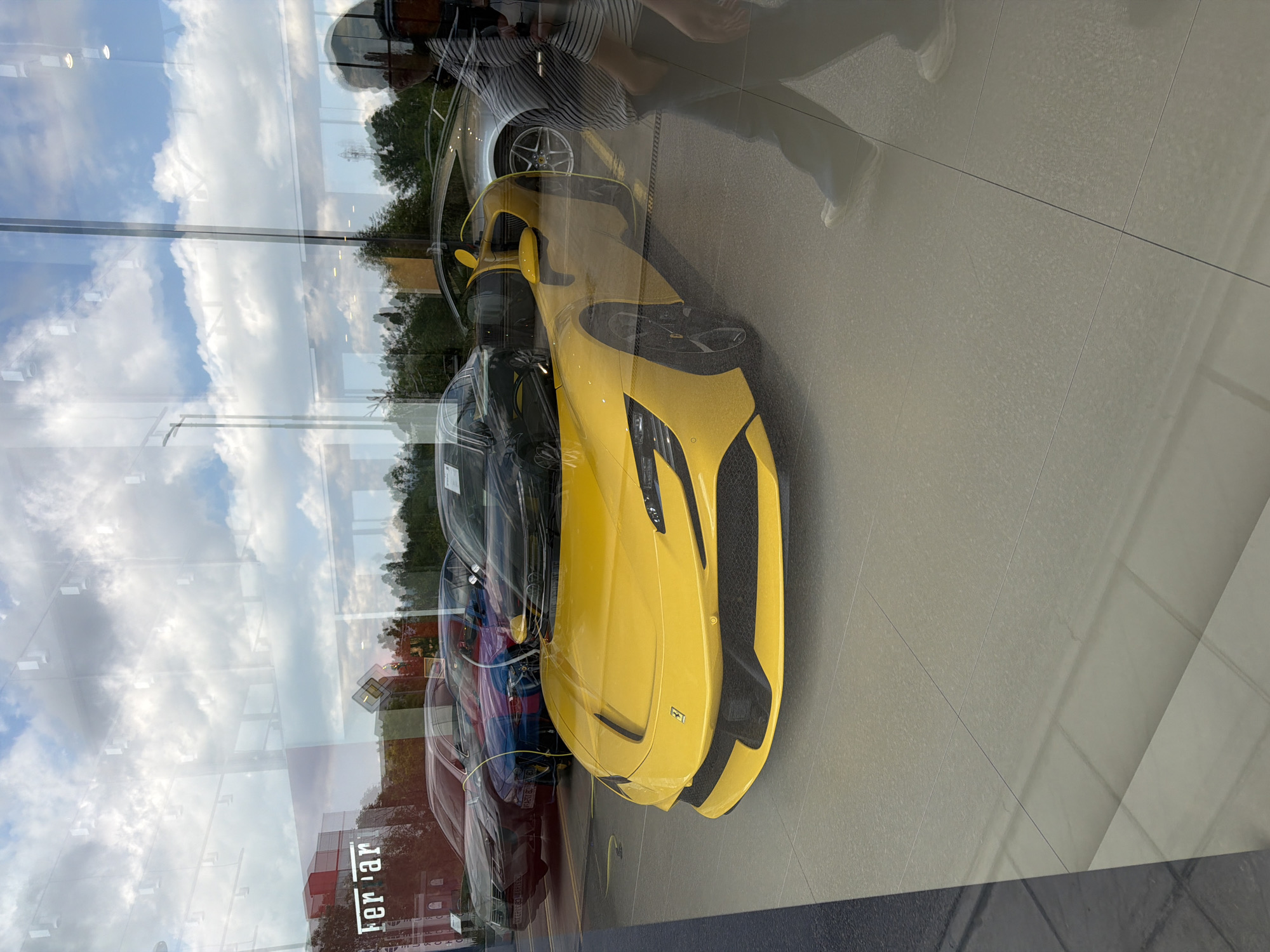 The image, taken through a large reflective glass window, captures a car dealership showroom with prominent external reflections. The primary subject is a bright yellow Ferrari sports car, sleek and modern in design, positioned in the foreground inside the showroom. Its front features black grilles and a small Ferrari emblem on the hood. Behind it, a silver car is partially visible, and further back, other vehicles, including shades of dark blue and red, are lined up. The showroom floor is made of light-colored tiles.
The activity depicted is an observation of the cars from outside the dealership. This perspective results in significant reflections on the glass. A cloudy blue sky, green trees, and distant buildings are clearly mirrored, overlapping the view of the interior. A person wearing a striped shirt and dark pants is reflected in the upper right portion of the glass, seemingly looking into the showroom.
It is daytime, indicated by the natural light and the bright sky in the reflections. The weather appears partly cloudy. A notable detail is the layering effect created by the transparent glass allowing a view of the showroom and the simultaneous strong reflections of the outside world. The word "Ferrari" is visible in white, reversed and partially cropped, as a reflection on the lower left side of the glass.