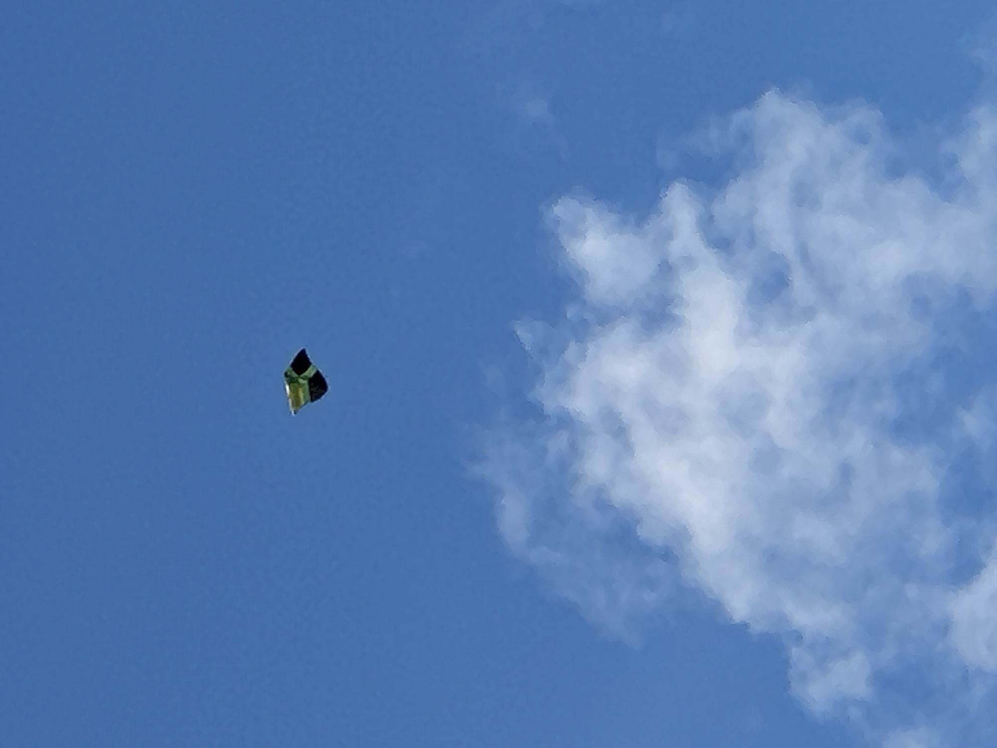 A small, colorful kite with green and black panels is airborne against a bright blue sky. Below the kite, large, fluffy white clouds drift across the sky. The kite appears to be in the upper left quadrant of the image, and the clouds occupy the lower half. The scene is outdoors and appears to be during the daytime.