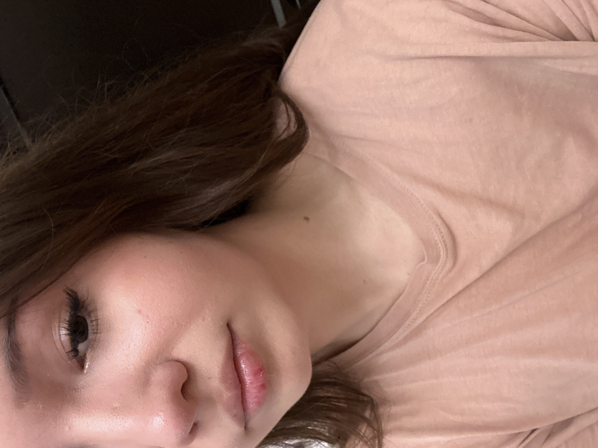The image is a close-up portrait of a person, likely female, captured from the chest up, with a dark, blurred background. The individual's head is tilted, showing part of their face and upper body at an angle. They have long, dark brown hair that cascades around their head and shoulders.
The person is wearing a light peach or pink-colored t-shirt with visible fabric texture and subtle folds. Their right eye is visible, featuring dark eyelashes and a dark iris. The skin on the face appears natural, showing some minor redness near the nose and a healthy glow. The lips are slightly parted, and the lower lip exhibits some redness and what appears to be a small area of irritation or chapping. A small, dark mole is visible on the neck/shoulder area, just above the t-shirt.
The overall setting appears to be indoors, possibly with low or ambient lighting, contributing to the soft, somewhat intimate feel of the image. The expression is neutral and relaxed. No specific activity is clearly depicted other than posing for the portrait. There is no visible text in the image. The image is associated with Naberezhnye Chelny, Russia.