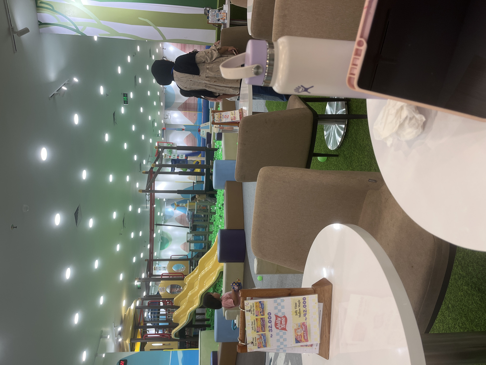 The image depicts an indoor playground with a ball pit and a yellow slide. In the foreground, there are two round white tables, one of which has a crumpled tissue and a pink-cased electronic device. A light purple water bottle with a black lid and a Hydro Flask logo is visible on the table. Two tan chairs are placed around the table. To the left, a wooden stand displays several promotional flyers for "Jolly Time" products, with prices indicated in Indonesian Rupiah. In the background, a young child in a pink shirt plays near the ball pit. A woman wearing a black hijab and a patterned beige top stands near the playground. The environment appears to be a family entertainment center or play area. The presence of "Jolly Time" branding, a popular snack brand in Indonesia, and the general aesthetic suggest this location could be within a mall in Bandung City, Indonesia.
