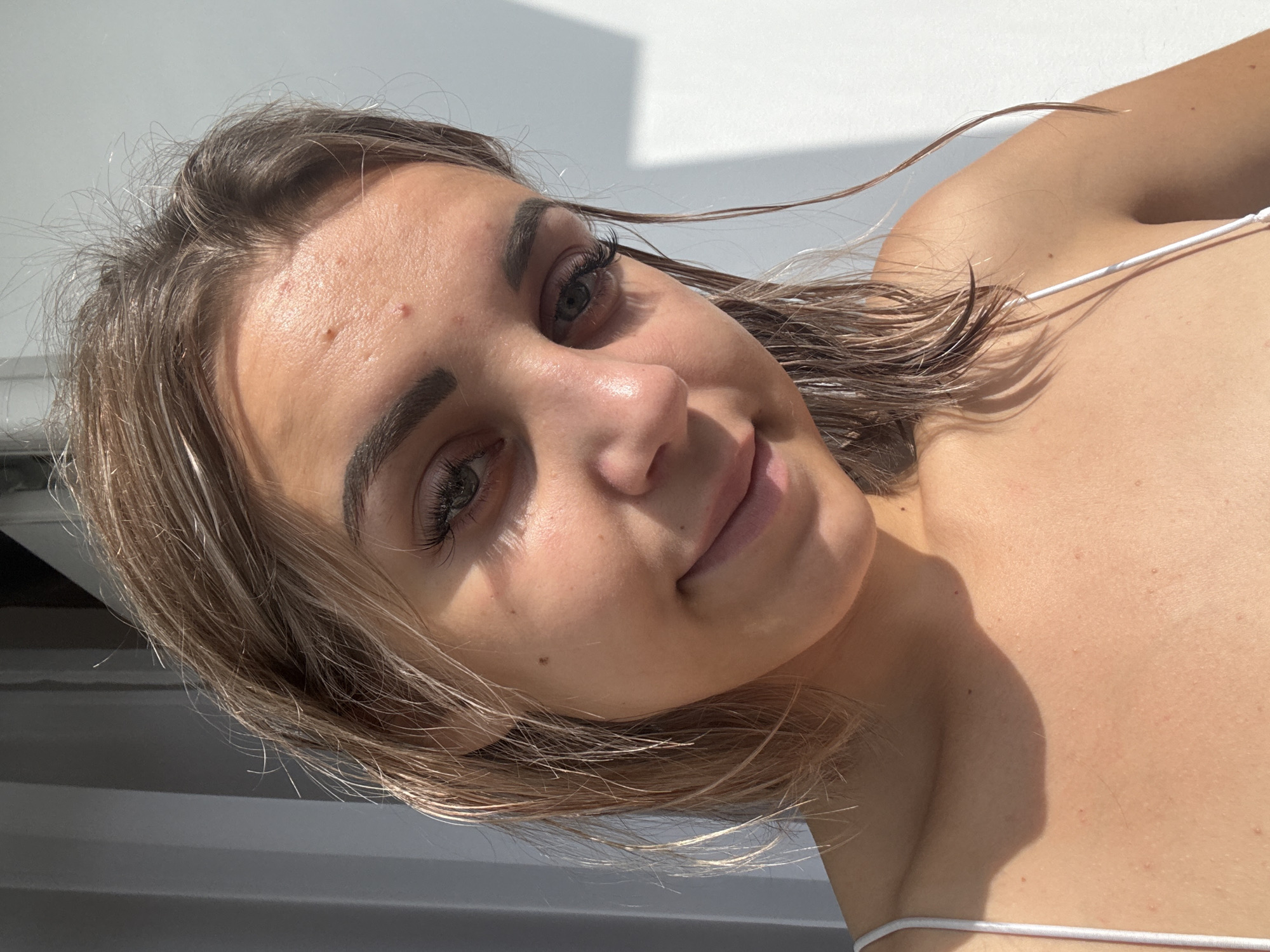 This is a close-up, high-angle portrait of a young Caucasian woman with fair skin, light brown hair, and green eyes. She is facing the camera, slightly angled to her right, and smiling subtly. Her hair is wet and reaches just past her shoulders, with some strands falling across her face. She has noticeable eyelashes and eyebrows. Her forehead shows a few small blemishes. She is wearing a white tank top with thin straps. The lighting suggests a sunny environment, casting shadows on her face and shoulder. The background is a plain white wall with a soft shadow pattern. The image does not contain any specific landmarks or elements that would indicate a location in Kholmsk, Russia, or any city.