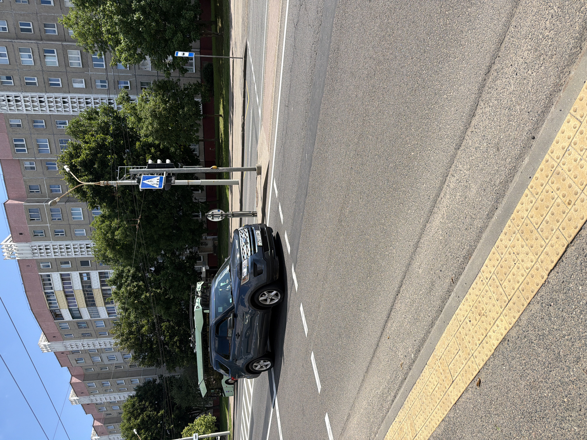 The image shows a street scene in Minsk, Belarus. In the foreground, a grey Ford Explorer SUV is driving on a paved road. The vehicle has a license plate that reads "2571 XM-7". White dashed lines mark a pedestrian crossing on the road. To the left of the road, a textured yellow tactile paving surface is visible. Behind the car, a green trolleybus is parked. In the background, a tall, multi-story apartment building with pink and grey facades stands against a clear blue sky. Trees line the area between the road and the building. A traffic light and pedestrian crossing signs are also present.