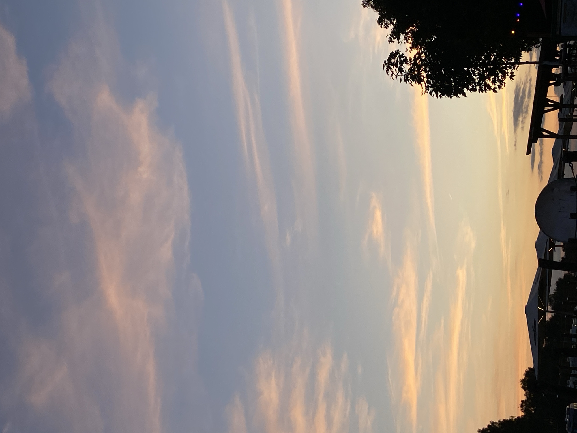 The image depicts a twilight sky over Rinteln, Germany. The sky is a gradient of pale blue and pink, with wispy clouds catching the last rays of the sun. Silhouetted against this vibrant sky are the tops of trees and some man-made structures, possibly part of a beer garden or outdoor seating area, including several large, white umbrella-like canopies and a wooden overhang. The scene is calm and serene, suggesting the end of a day. There are no people clearly visible in the image.