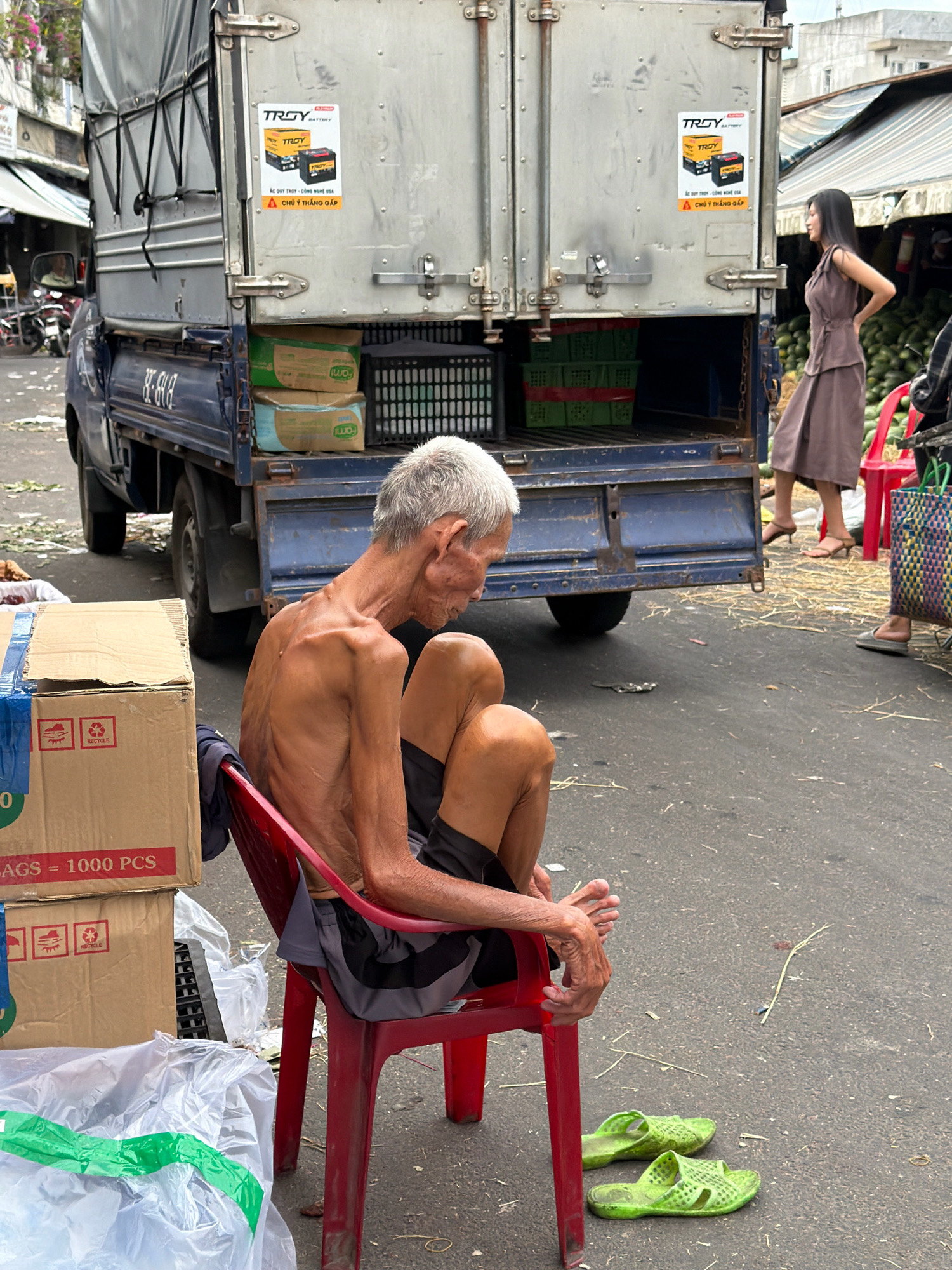 The image depicts a street scene in Nha Trang, Vietnam, likely near a market or distribution hub, given the presence of a delivery truck and stacked boxes. In the foreground, an elderly, emaciated man with grey hair is seated on a red plastic chair, wearing dark shorts and a t-shirt, his upper body bare. His posture suggests weariness or rest. To his left are several cardboard boxes, one prominently displaying "Bags = 1000 PCS" and recycling symbols. To his right, a pair of bright green plastic sandals are placed on the asphalt. In the midground, a large blue delivery truck with a grey cargo container is parked. Stickers advertising "TROY BATTERY" are visible on the back of the truck. The truck bed contains green crates and more boxes. A woman in a brown dress is standing to the right of the truck, looking away from the camera. The environment is an outdoor urban setting with a paved road surface, some scattered debris, and the suggestion of market stalls or buildings in the background.