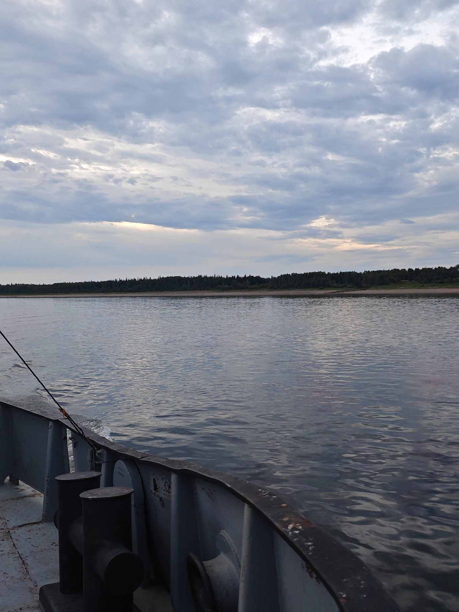 This media file depicts a view from a boat on a body of water, likely a river, with a tree-lined shore in the distance. The foreground shows the deck of a boat, with metal railings and two bollards. A dark rope is visible, extending from the boat into the water. The water itself is calm with ripples, reflecting the cloudy sky. The background features a dense forest along a sandy shore, typical of a northern landscape.
The sky is overcast with dramatic clouds, suggesting it might be late afternoon or early evening, or possibly a cool, cloudy day. The lighting is subdued, creating a muted color palette. The overall mood is serene and contemplative, with a sense of solitude and the vastness of nature. There are no people visible in the image. The location is identified as Nikulino, Russia, which aligns with the description of a forested shore and river landscape. There is no visible text in the image.