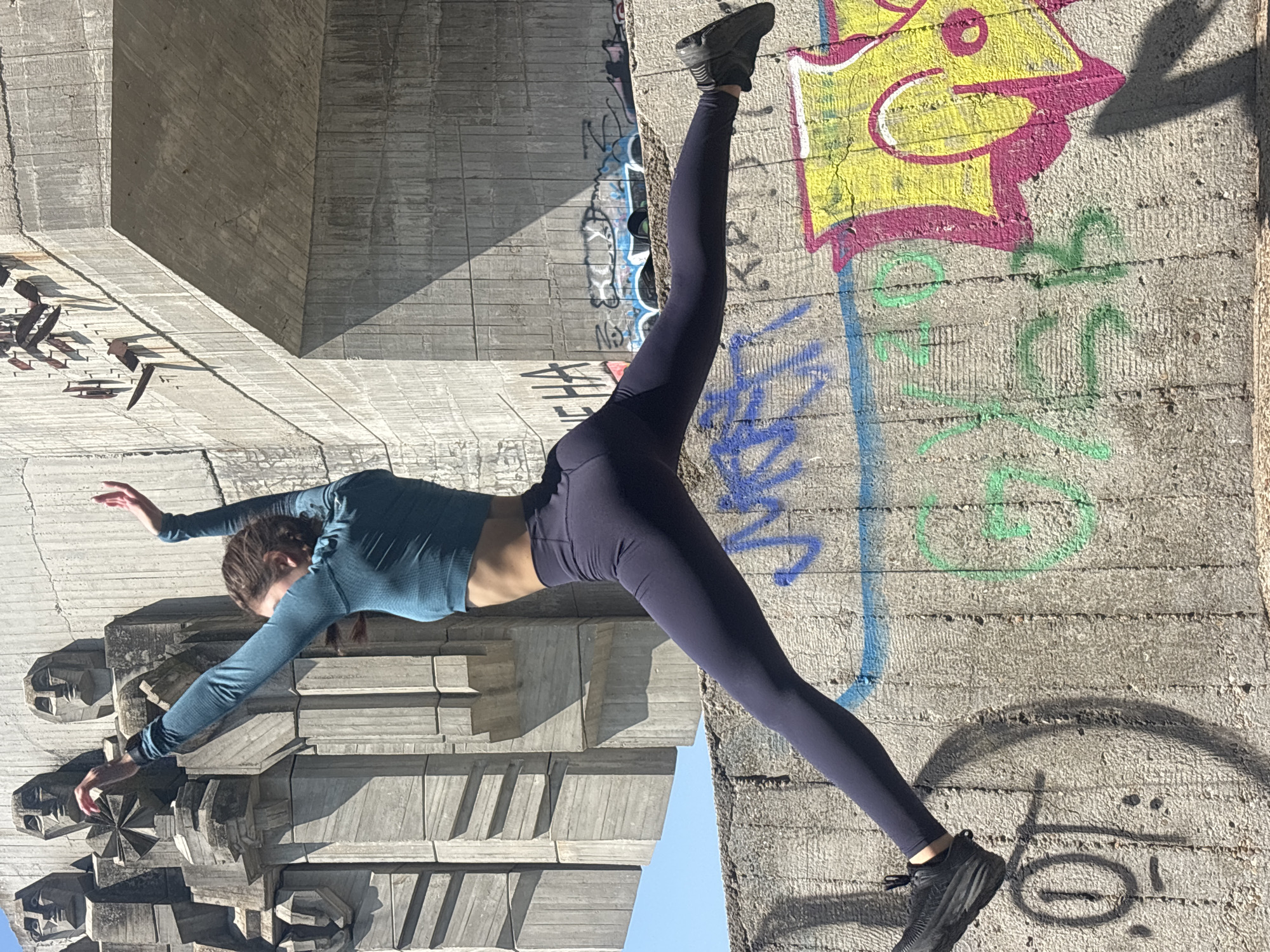 An outdoor, eye-level, full-shot image captures a female individual performing a dynamic split jump against a brutalist architectural backdrop in Varna, Bulgaria. The subject, oriented horizontally across the lower and mid-frame, wears a teal long-sleeved crop top, dark navy athletic leggings, and black athletic shoes. Her arms are raised and bent at the elbows with open hands, and her head is turned to her right.
The background on the left features a portion of the Pantheon of the Fallen Fighters Against Fascism and Capitalism, a monument characterized by large, textured concrete sculptures of stylized human heads and torsos. To the right, a massive concrete bridge or overpass structure is visible, with sharp angles and a raw, textured finish, creating a deep shadow beneath its span.
The immediate foreground is dominated by a rough-textured concrete wall extensively covered in colorful graffiti. Visible markings include blue stylized text and squiggles, green letters, and vibrant yellow and magenta bubble letters. A large, black-outlined smiley face with a dash for a nose is partially visible on the left side of the wall.
The scene is under bright sunlight, evidenced by the sharp, defined shadows cast by the architectural elements and the subject, particularly the shadow of her left leg and foot on the graffiti-laden wall. A clear, light blue sky is visible in the upper left quadrant.