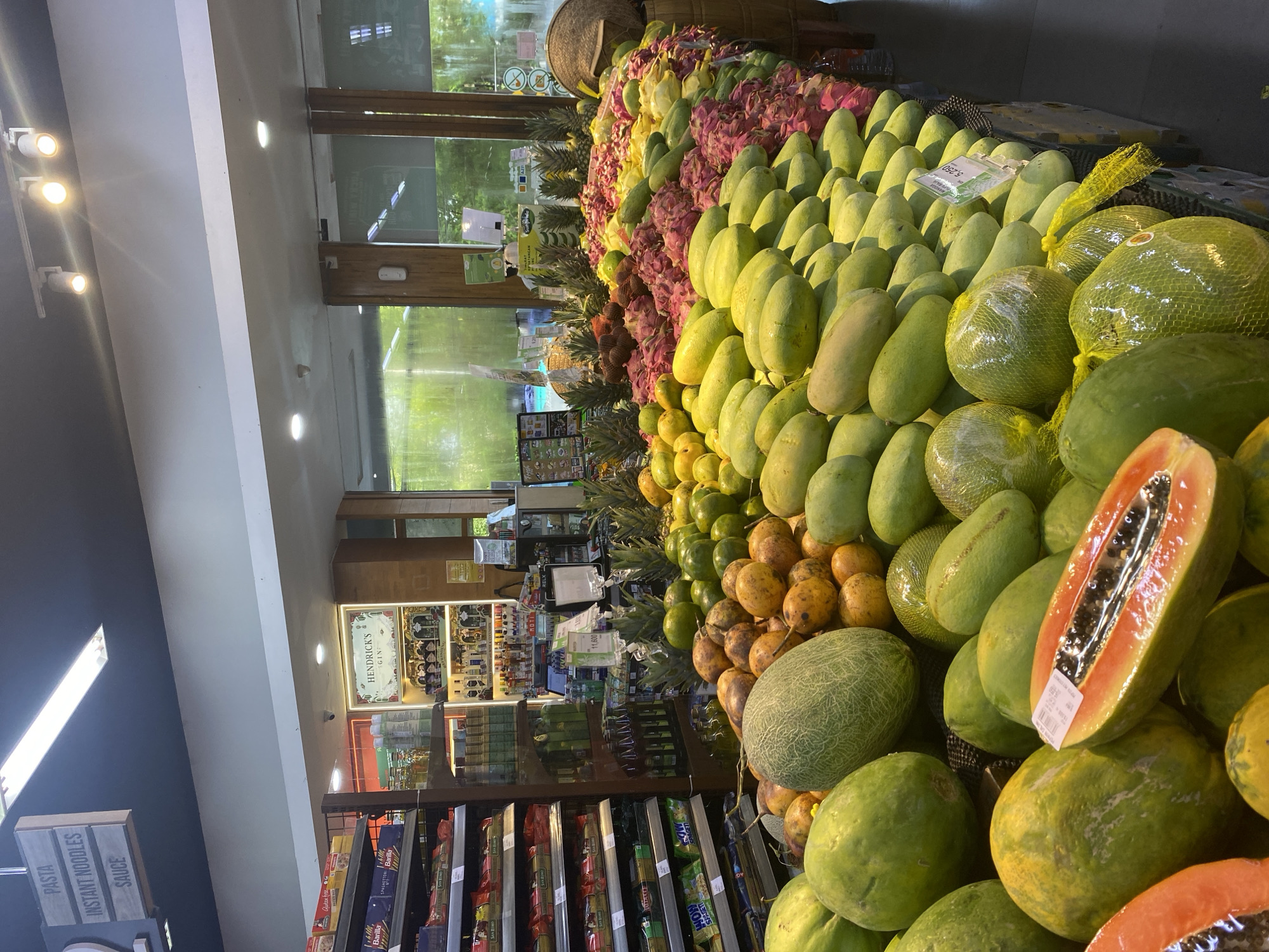 The image depicts a grocery store display of fruits and vegetables, situated in North Kuta, Indonesia. A large pile of produce including mangoes, dragon fruits, pineapples, melons, and papayas are arranged attractively. Shelves stocked with pasta, sauces, and other grocery items are visible in the background. A sign for Hendrick's Gin is also visible. The store appears well-lit, with natural light coming through the window, and is tidy.