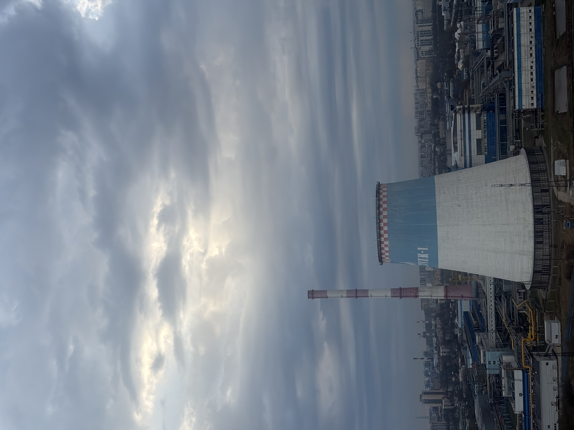 The image presents an industrial landscape dominated by a large cooling tower and a tall smokestack, set against a cloudy sky and a distant cityscape. No people are visible.
The scene depicts an active industrial facility, likely a thermal power plant or combined heat and power (CHP) plant, given the presence of the cooling tower and smokestack. No specific activity or event is captured; it's a static overview of the infrastructure.
It is daytime, characterized by an overcast sky with heavy, grey clouds, some areas appearing brighter where sunlight partially penetrates. The overall light is muted. The prominent cooling tower features a light blue and white color scheme, topped with a distinctive red and white checkered pattern around its rim. Adjacent to it, a slender smokestack rises, painted with alternating red and white horizontal stripes. The industrial complex below consists of various buildings, pipes, and structures, rendered in shades of blue, white, and grey. In the background, a sprawling urban area with numerous multi-story buildings extends to the horizon, suggesting a location at the edge of a city.
On the blue section of the large cooling tower, the Cyrillic text "ТЭЦ-1" is clearly visible, which translates to "TPP-1" or "CHP-1" (Thermal Power Plant No. 1 or Combined Heat and Power Plant No. 1).