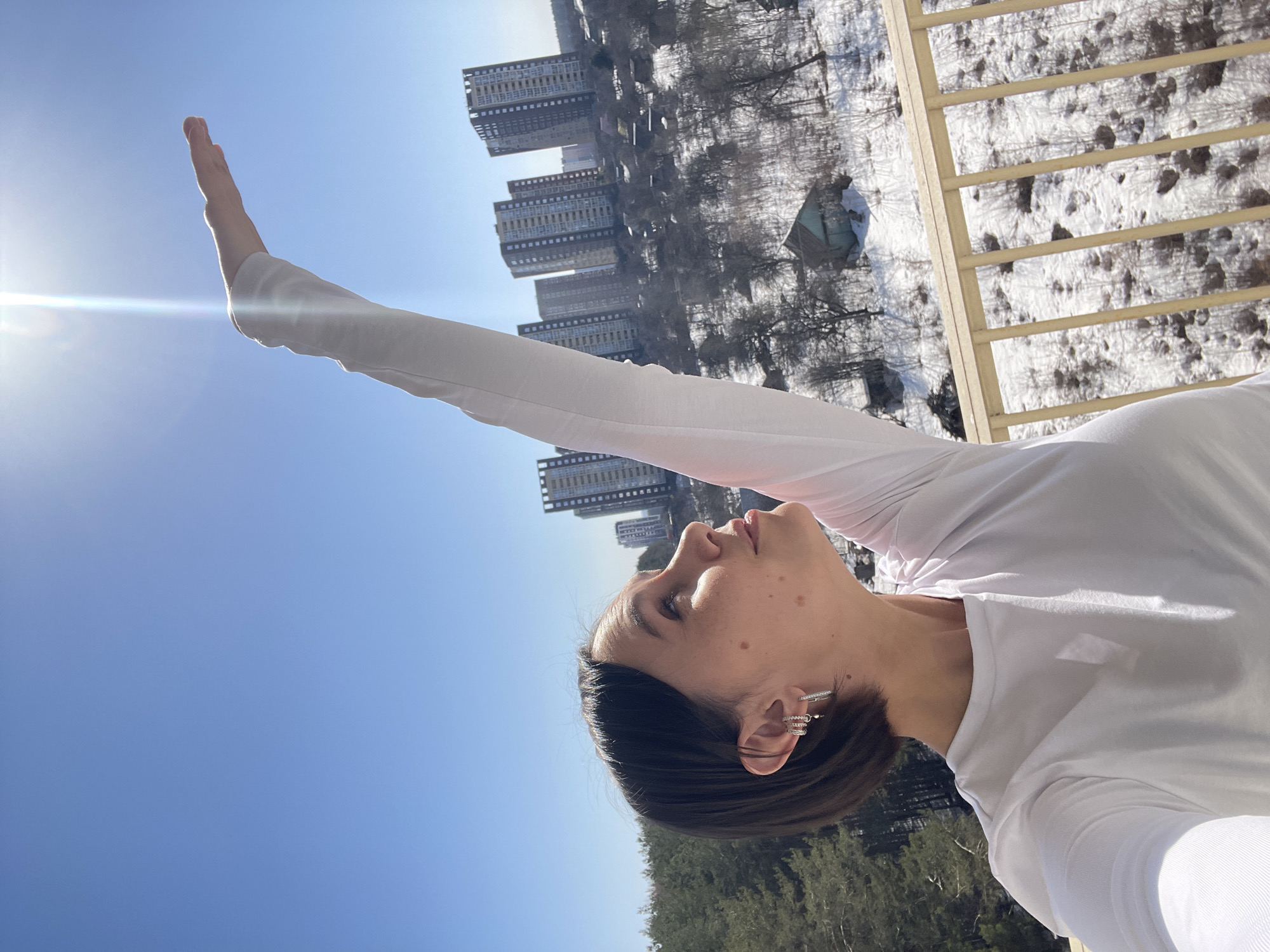 A woman with dark hair is taking a selfie outside on a sunny day. She is wearing a white long-sleeved shirt and multiple earrings. Her right arm is extended upward, toward the sun. Behind her, multiple high-rise apartment buildings are visible. In the background, there are snowy areas, trees, and a wooden railing. Based on the apartment buildings and landscape, the location is Izhevsk, Russia.