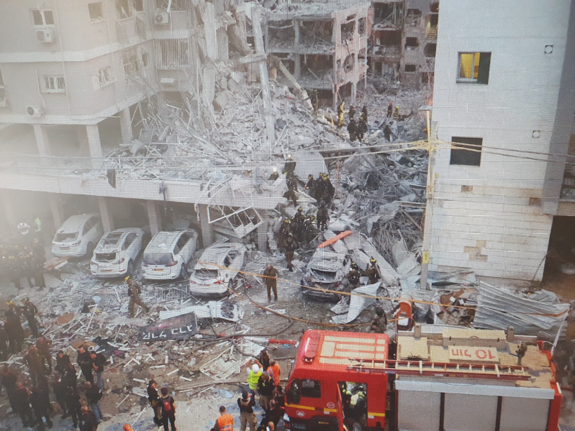The image depicts the aftermath of a significant structural collapse, likely an explosion, in an urban environment. A multi-story building is severely damaged, with its facade blown out and debris scattered across a wide area. The collapsed section reveals the internal structure of the building, with twisted metal and concrete.
In the foreground, several white cars are parked, some of which appear to be damaged by the debris. A red fire truck is visible, with its ladder extended. Numerous individuals, many wearing protective gear such as helmets and vests, are present at the scene. They appear to be emergency responders, possibly involved in search and rescue operations, as well as onlookers. Some individuals are standing in groups, while others are moving through the debris field. Yellow caution tape is present, cordoning off sections of the area.
The environment appears to be a residential or mixed-use area, given the presence of cars and the multi-story buildings. The sky is not visible in the frame, suggesting an elevated viewpoint or dense urban surroundings. The image does not contain any specific indicators of its location within Tashkent, Uzbekistan, or any other city.