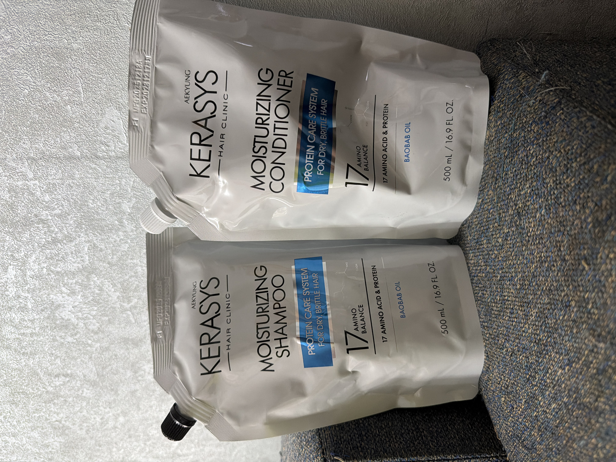 Two pouches of Kerasys brand hair care products, labeled "Moisturizing Shampoo" and "Moisturizing Conditioner", are placed side-by-side on a textured, dark blue and grey upholstered surface, possibly a sofa or chair. The background is a textured, light-colored wall. Both pouches are white with black and blue text and graphics. The left pouch is slightly closer to the viewer and features the product name "MOISTURIZING SHAMPOO". The right pouch is slightly further away and displays "MOISTURIZING CONDITIONER". Both pouches indicate "AEKYUNG HAIR CLINIC" and highlight "PROTEIN CARE SYSTEM FOR DRY, BRITTLE HAIR" with "17 AMINO BALANCE" and "17 AMINO ACID & PROTEIN" information. The volume is stated as "500 mL / 16.9 FL. OZ." on both. A black screw-top cap is visible on the left pouch. The scene is indoors, likely a domestic setting. The image does not contain any visual information that would suggest a specific location within Chaikovsky, Russia.