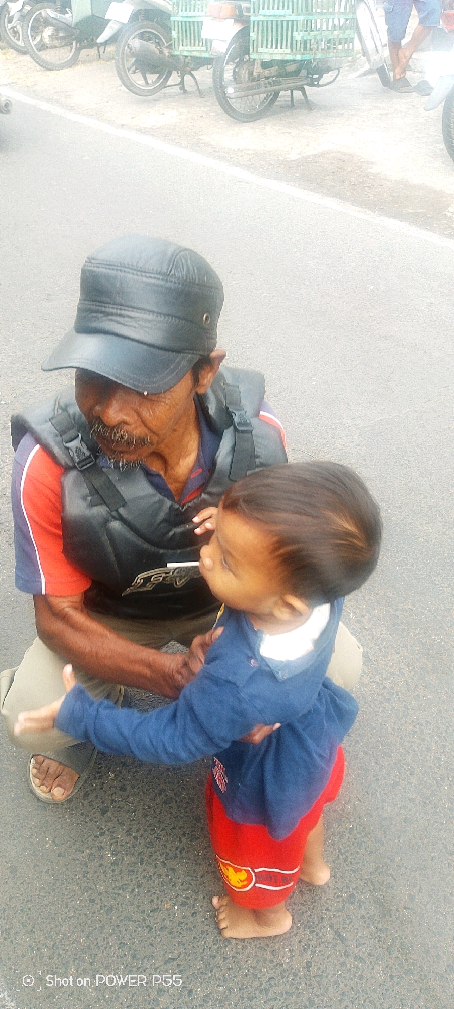An older man wearing a black cap and a black vest over a red and blue shirt is kneeling on an asphalt road. He is holding a young child who is wearing a blue long-sleeved shirt and red shorts. The child is being held close to the man and appears to be looking at a white object held between their lips. In the background, several motorcycles are parked, some of which have large cages attached to them. The environment appears to be a roadside in a city.