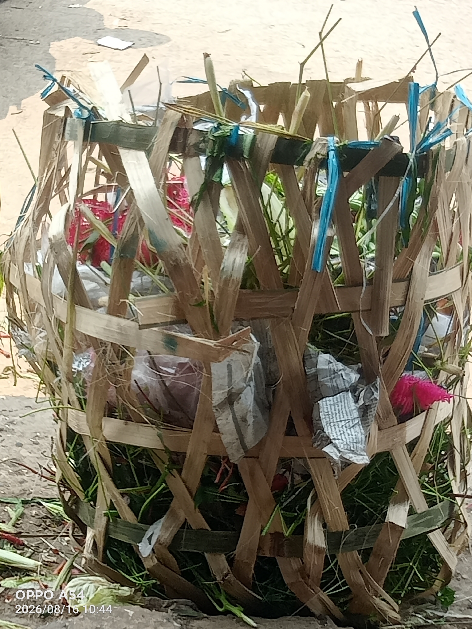 An outdoor, eye-level, close-up shot features a woven bamboo basket filled with green foliage, what appears to be pink flowers, and some crumpled paper. The basket is constructed from thin strips of bamboo woven in a crisscross pattern, with some blue twine used to bind sections together. The basket sits on a dirt or gravel surface, and debris, including more green foliage, is scattered around its base. The lighting suggests it is daytime. A watermark in the bottom left corner indicates the photo was taken with an "OPPO A54" on "2026/08/16 10:44". While the image is tagged as being in Jambi, Indonesia, there are no specific visual cues within the image itself to confirm this location.