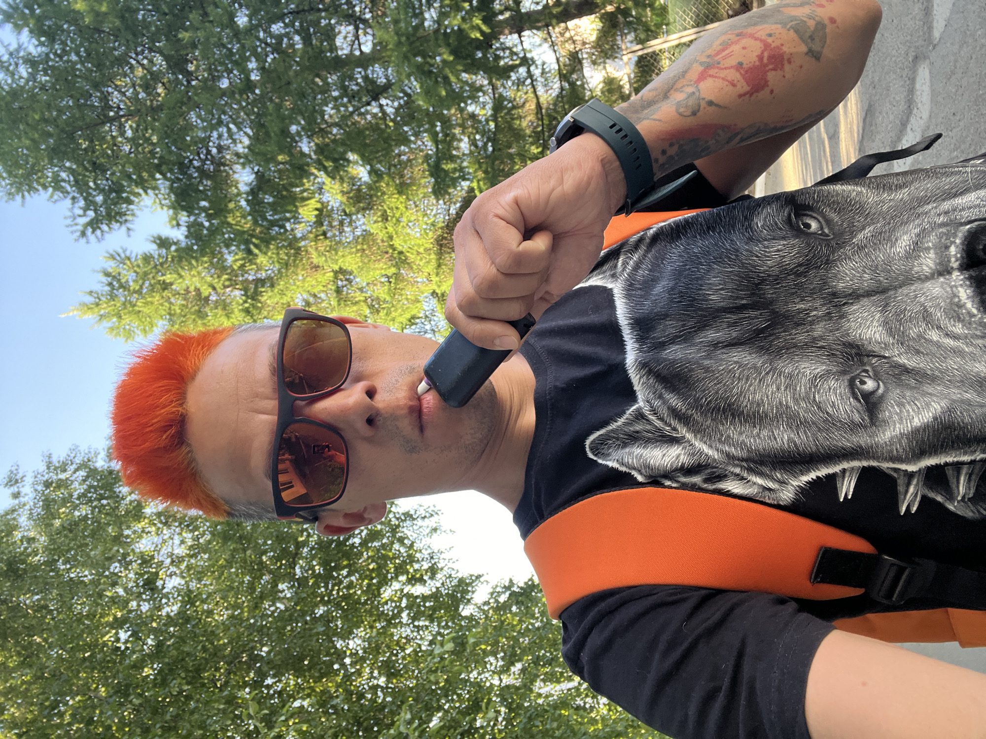 A man is outdoors, likely in a park setting with trees overhead. He has bright orange hair, sunglasses, and a black shirt. He is holding a vape device to his mouth. He is wearing a backpack with an image of a gray wolf's head on it and orange straps. He has a dark-colored smartwatch on his left wrist and a tattoo on his left arm. The picture's perspective is from below, looking up at the man. There are no clear indicators that this is in Chaikovsky, Russia.