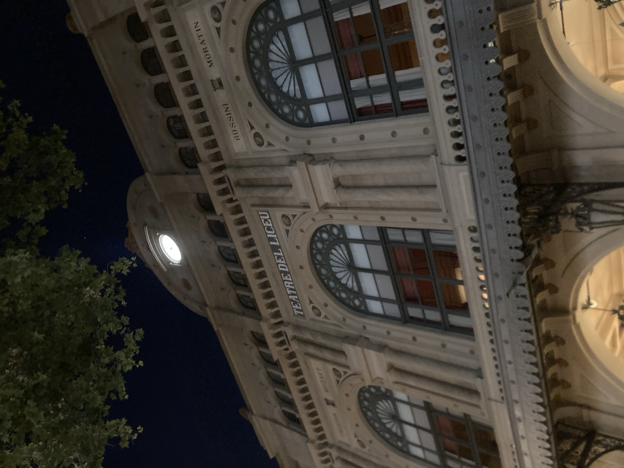 This is a nighttime, low-angle shot of the exterior of the Gran Teatre del Liceu opera house in Barcelona, Spain. The building's ornate facade, characterized by its elaborate stone carvings, decorative moldings, and arched windows, is illuminated. The windows, with their dark frames and subtle red curtains visible within, are a prominent feature, some with decorative fanlight designs above them.
The image captures a section of the building's upper levels, showcasing classical architectural elements. Above some of the windows, text is visible in capital letters. To the left of the image, a portion of a leafy green tree is visible against a dark night sky. A circular illuminated clock face is also visible on the building's facade. The overall impression is one of grand architecture set against the night sky, likely in a city environment. The lighting suggests the opera house is either open for an evening performance or has exterior lighting.