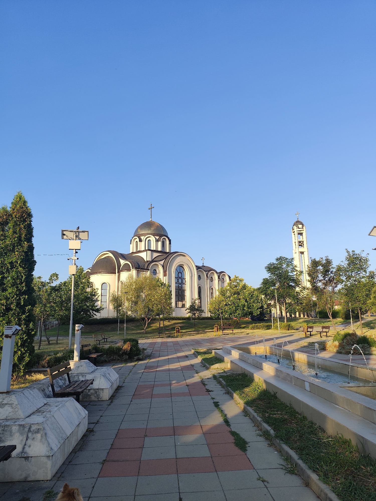 The image shows a view of the "St. Patriarch Evtimiy" Orthodox Church and its surroundings in the city of Montana, Bulgaria. The church, characterized by its white facade, multiple domes topped with crosses, and arched windows, is the central subject. To the right of the main church building, a tall bell tower with arches and bells is visible.
In the foreground, a paved walkway with a checkered pattern of reddish-brown and grey tiles leads towards the church. On either side of the walkway are decorative stone benches with wooden seating. A multi-tiered water fountain with several jets of water is also a prominent feature on the right side of the walkway, surrounded by grass.
The area around the church and walkway is landscaped with various trees, including tall evergreens on the left and deciduous trees with green foliage. Benches are scattered throughout the park-like setting. The sky is clear and blue, indicating fair weather. A small dog is visible in the lower left corner of the image. A street lamp with what appears to be a bird perched on it is also situated to the left of the main church structure.