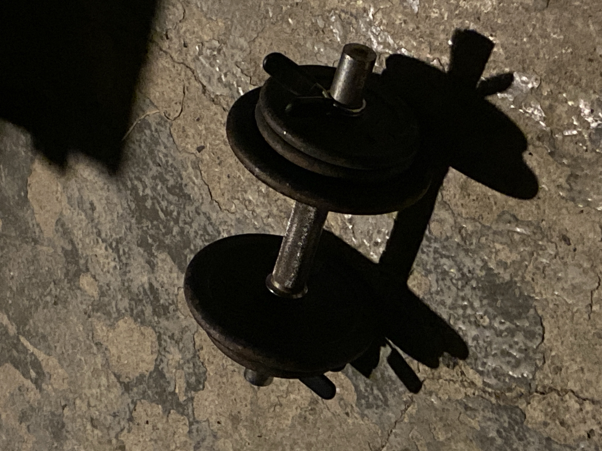 A dumbbell rests on a cracked, uneven concrete surface. The dumbbell is partially obscured by shadow. The location is not discernable from the image. No people are visible.