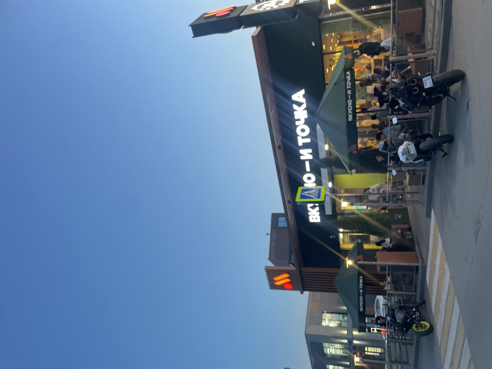 This image shows a Вкусно — и точка restaurant in Kazan, Russia, with outdoor seating where people are gathered. There are motorcycles parked nearby. The sky is a clear blue, and the restaurant is situated near other buildings. The Вкусно — и точка branding is visible on the signage and awnings. A pedestrian crossing is in the foreground.