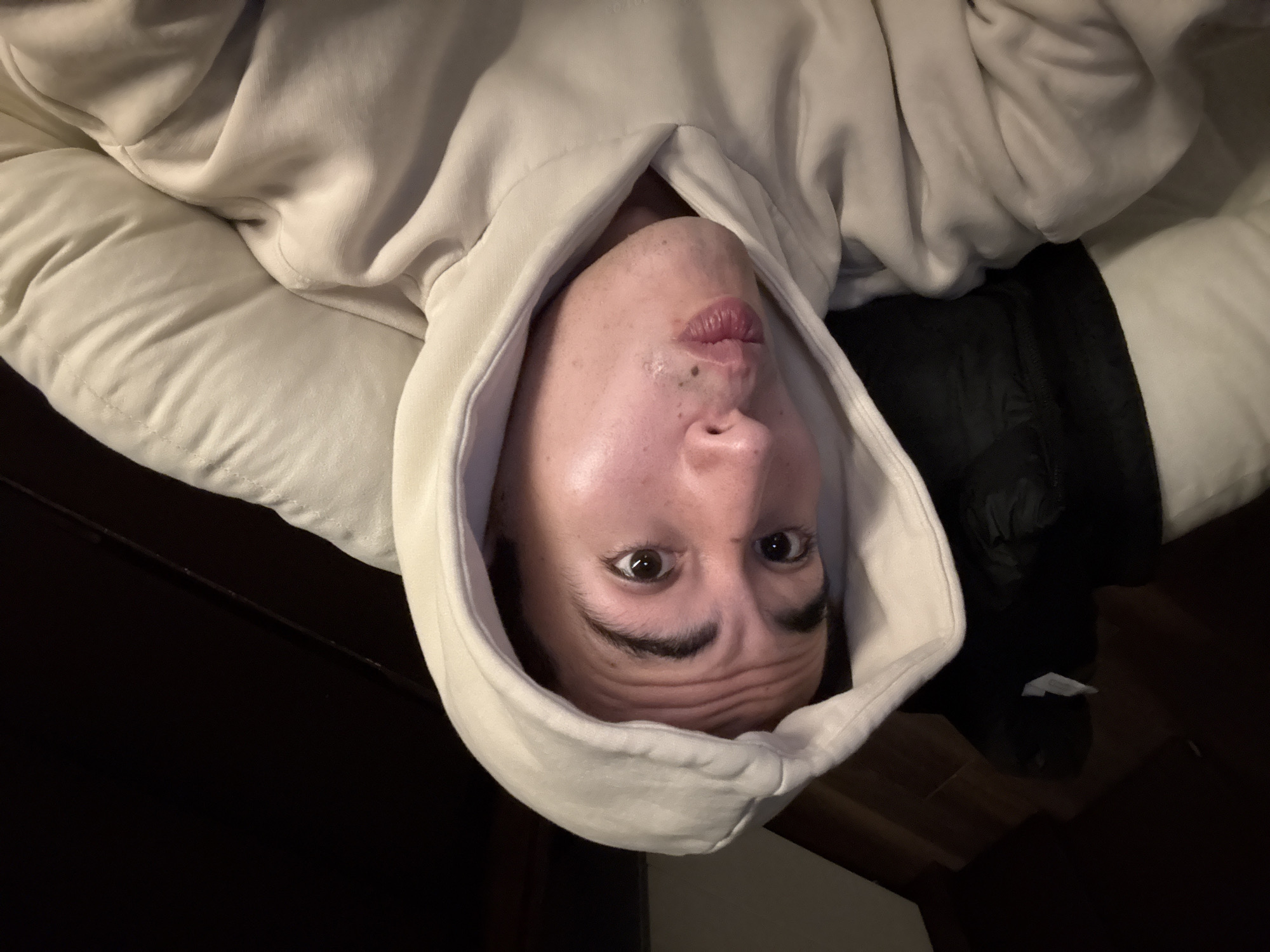 The image is an inverted portrait showing a person lying down, their head at the bottom of the frame and their feet (unseen) towards the top, implying the camera was upside down or the image was rotated 180 degrees. The subject is a person, likely young adult, wearing a light-colored (off-white or cream) hooded sweatshirt, with the hood framing their face. They are positioned against a soft, light-colored surface, possibly a pillow or cushion.
The person looks directly at the camera with wide, expressive eyes and dark, well-defined eyebrows. Their mouth is puckered in a "fish face" expression, and their forehead shows several horizontal wrinkles, contributing to a playful or surprised look. A few small, dark moles or marks are visible on their upper lip/chin area. Their skin has a slight sheen, suggesting indoor artificial lighting.
To the right of the person's head, a crumpled dark fabric or item of clothing is visible. The background consists of dark tones, including what appears to be dark furniture or wooden floorboards and steps, with a thin dark cable and a small white tag or object faintly visible on the dark surfaces. The overall scene suggests an indoor, casual setting, possibly a bedroom or living room, capturing a lighthearted moment. There is no visible text. The inferred location is San Bernardino Tlaxcalancingo, Mexico.