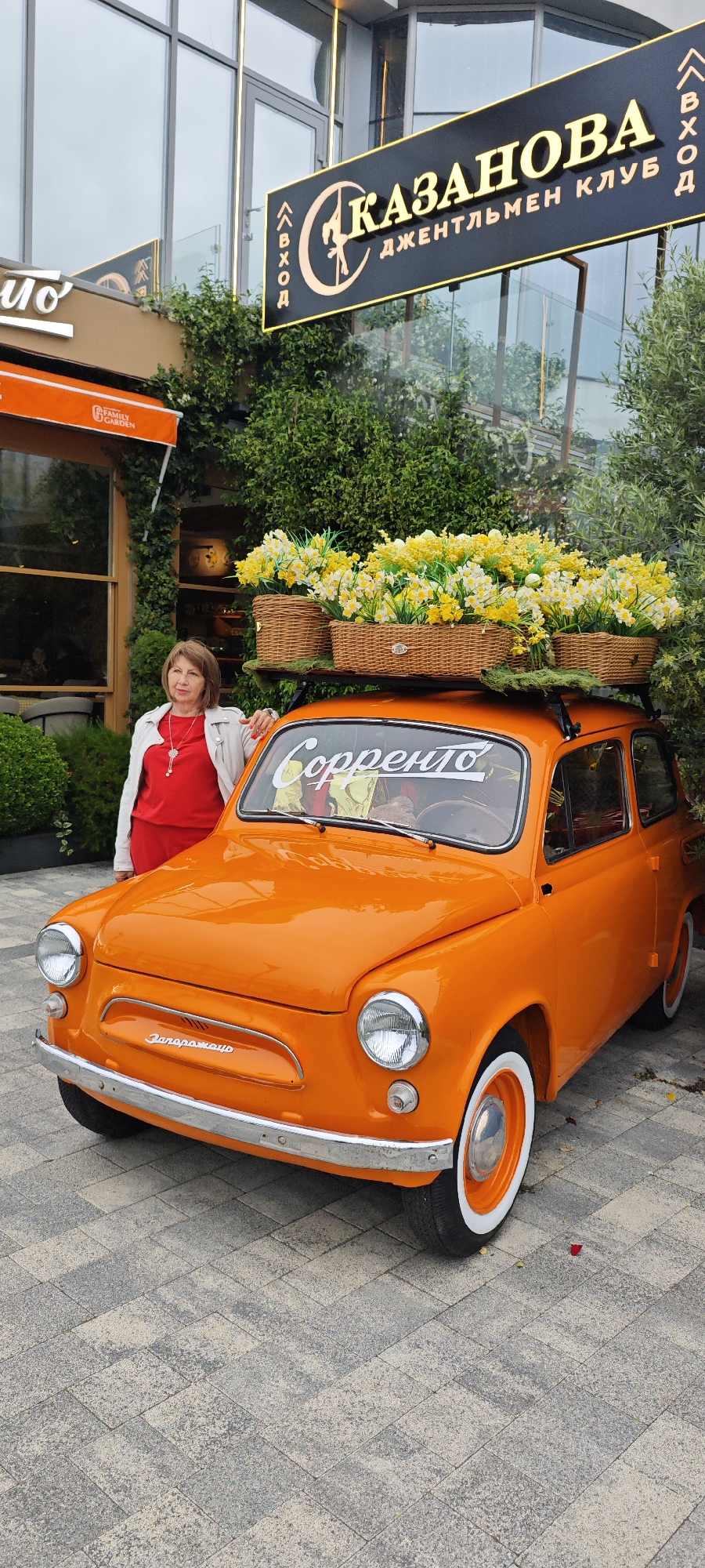 An eye-level, full outdoor shot depicts an orange Zaporozhets car adorned with flowers, standing in front of a building in Sochi, Russia. A woman stands next to the car, leaning her left hand on the hood and facing the camera.
The orange Zaporozhets car is the focal point, with four wicker baskets filled with yellow and white flowers on its roof rack. A white sign with "Соppento" is visible on the windshield. The car has classic whitewall tires and chrome detailing on the bumper and around the headlights.
The woman on the left is dressed in a red top and wears a white jacket. She has a necklace with a pendant. She is positioned near the driver's side, partly obscured by the car.
Behind the car is a building with green foliage and a sign that reads "КАЗАНОВА ДЖЕНТЛЬМЕН КЛУБ," indicating "Kazanova Gentlemen's Club." The entrance door is visible to the left, with orange awnings above.
The ground is paved with grey rectangular stones. The sky is overcast, and the image is well-lit, but the light is flat. The setting appears to be a street or public area in Sochi, Russia, as identified by the Cyrillic script and architectural style.