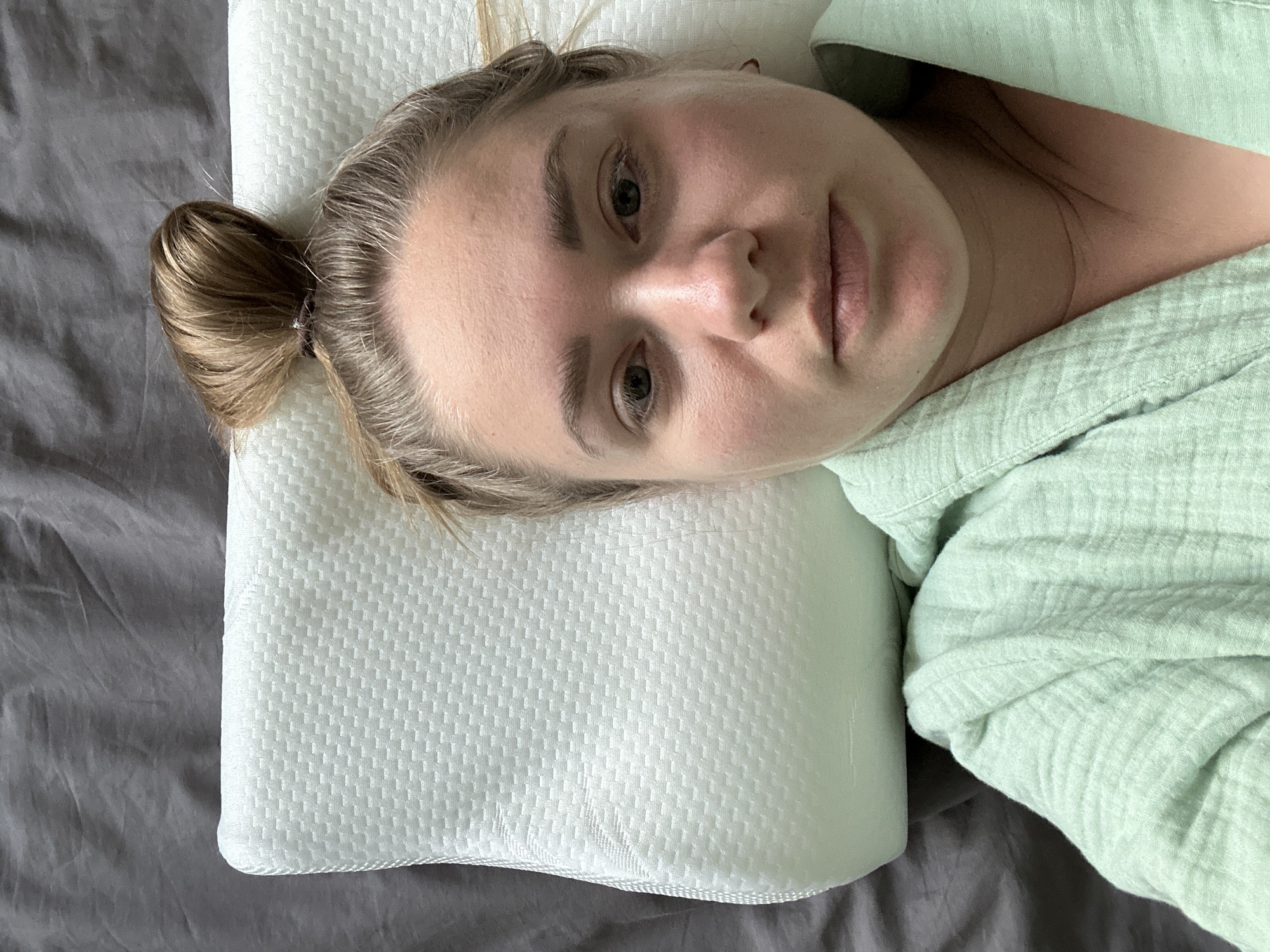 An overhead photograph depicts a single female individual lying on her back. Her head rests on a light-colored, contoured ergonomic pillow with a textured, grid-patterned cover. Dark gray, wrinkled bedding is visible behind and to the left of the pillow. The woman has light brown hair pulled back into a messy bun on the crown of her head. She is wearing a light green, textured fabric garment, possibly a robe, visible around her neck and upper torso. Her face is directly centered in the frame, showing a neutral expression, with her eyes open and directed towards the camera. Her skin appears pale with some natural redness on her cheeks and nose. No other objects or individuals are present. The specific location in Chaikovsky, Russia, is not discernible from the image.