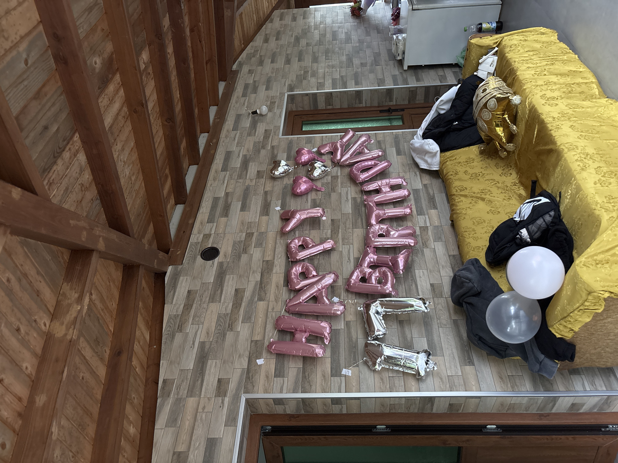 The image shows an interior view, likely in Zlatograd, Bulgaria, featuring a floor with faux wood tile arranged with "Happy Birthday" foil balloons. A light-colored sofa is on the right side, partially covered with clothing items and two round balloons, one white and one transparent gray. A gold decorative balloon is on the sofa. Along the upper right is a white refrigerator. A wooden ceiling structure is visible on the left. A door is visible at the bottom, and a window in the center. The scene appears to be set up for a birthday celebration.