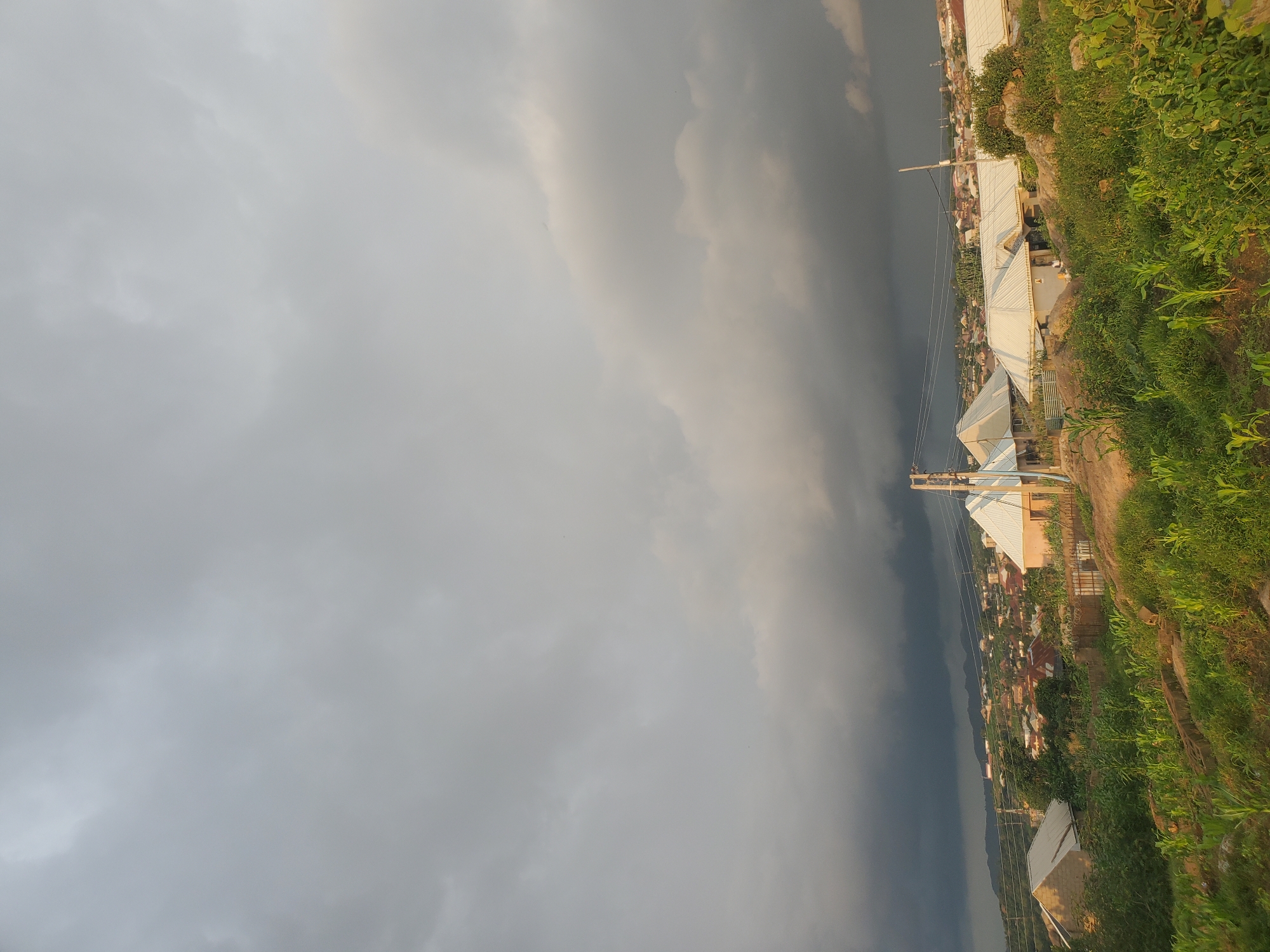 An eye-level, medium shot captures a residential area on a hillside in Jos, Nigeria, under a dramatic, overcast sky. The foreground is dominated by lush green vegetation, including what appears to be young corn stalks and other leafy plants, growing on a rocky incline. A dirt path or uneven ground is visible amidst the greenery.
Several houses with distinct white, gabled metal roofs are scattered across the mid-ground. Some of these houses are made of light-colored brick or plaster. A utility pole with wires stands to the right of center, and further in the distance, the town stretches across the landscape with numerous other buildings, their roofs varying in color, mostly reddish-brown.
The background is characterized by a brooding, dark gray cloud bank that looms over distant, hazy hills. The sky transitions from the dark, heavy clouds in the background to lighter, more diffused gray clouds overhead, suggesting an impending storm or recent rainfall. The overall lighting is dim and diffused, typical of a cloudy day. No people or specific actions are discernible in the image.