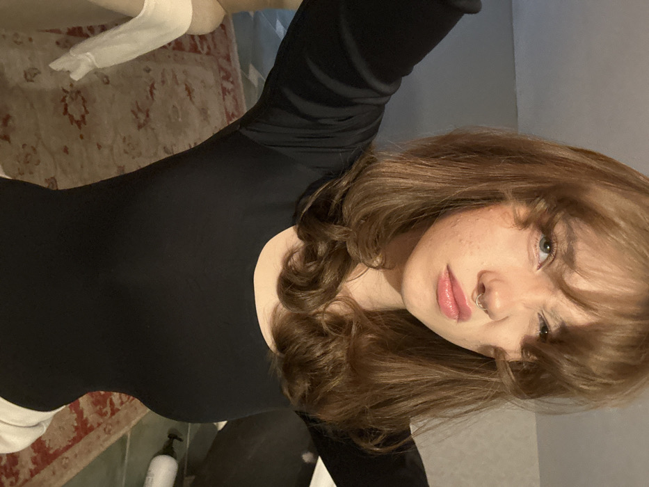 A single female subject is depicted in an indoor environment, presumed to be a room within a private residence in the city of Unknown, Unknown. The subject, a young adult with light brown, shoulder-length wavy hair and bangs, is positioned centrally, facing the camera directly. Her left eye is narrowed/squinted, and her mouth is slightly pursed; a silver septum piercing is visible. Minor dermatological blemishes are present on her cheeks. She is wearing a black, form-fitting, long-sleeved top with a scoop neck and light-colored lower garments, partially visible at the bottom of the frame. Her arms are raised and partially visible. The background consists of a patterned rug with red and cream hues covering the floor, a light-colored object resembling a mannequin limb or draped item to the left, and a white pump-dispenser bottle on a surface to the right. The walls are light gray/beige.