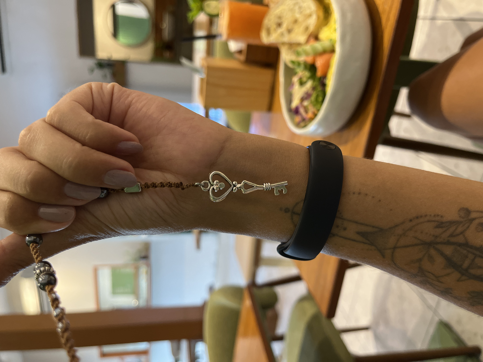 Here's a detailed description of the image:
**Scene:** The image appears to be taken indoors, likely in a restaurant or cafe setting. The foreground features a person's arm and hand, adorned with jewelry. A bowl of food is visible in the background, suggesting a meal setting.
**People:** Only a portion of one person is visible. Details about the person, besides their arm and hand, are not discernible.
**Subjects/Objects:**
* A person's arm and hand: The arm has a tattoo.
* Jewelry: A beaded bracelet with a key-shaped charm and a black fitness tracker are on the wrist.
* Food: A bowl containing a salad with various ingredients (possibly salmon, greens, bread) is in the background.
* Furniture: Wooden tables and chairs are partially visible.
* Plant/Decor: Some greenery is present in the background.
**Actions/Interactions:** The person is wearing the jewelry. It's implied they are at a table, possibly about to eat.
**Environment:** The location is identified as Kuta Selatan, Indonesia. However, there is no definitive element that allows it to be defined as being in the city of Kuta Selatan, Indonesia.
**Concise Description:** A person's arm wearing a beaded bracelet with a key charm and a black fitness tracker is in the foreground. A bowl of food is partially visible in the background in a restaurant setting. The location is identified as Kuta Selatan, Indonesia.