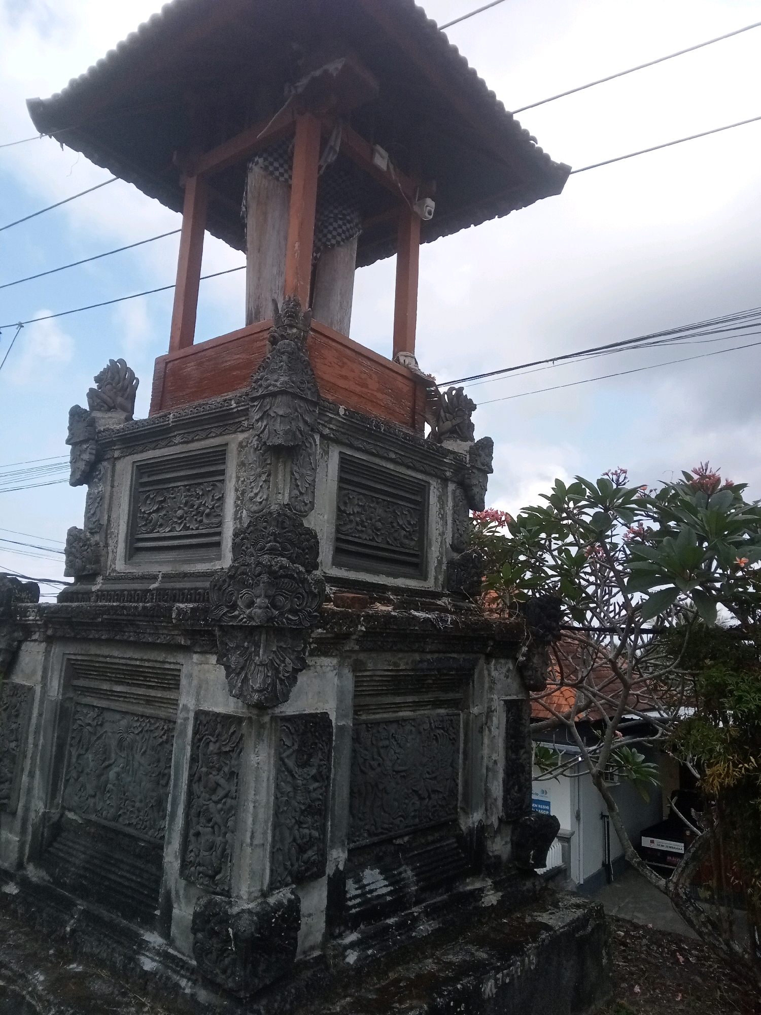The image depicts a traditional Balinese structure, likely a bell tower or shrine, with intricate stone carvings and a wooden roof. The structure is tall and weathered, suggesting it is an older building. In the background, there are power lines, a tree with pink flowers, and a building with some text visible on it, including "DEM JEMBRANA". The scene is set outdoors under a cloudy sky. Based on the text "DEM JEMBRANA" and the architectural style, the location is likely in the city of Mendoyo, Indonesia, which is part of the Jembrana Regency. There are no people visible in the image. The stone carvings on the base of the structure are detailed, featuring mythological figures and patterns. The wooden beams of the roof are visible, and the roof itself appears to be made of dark, corrugated material.
