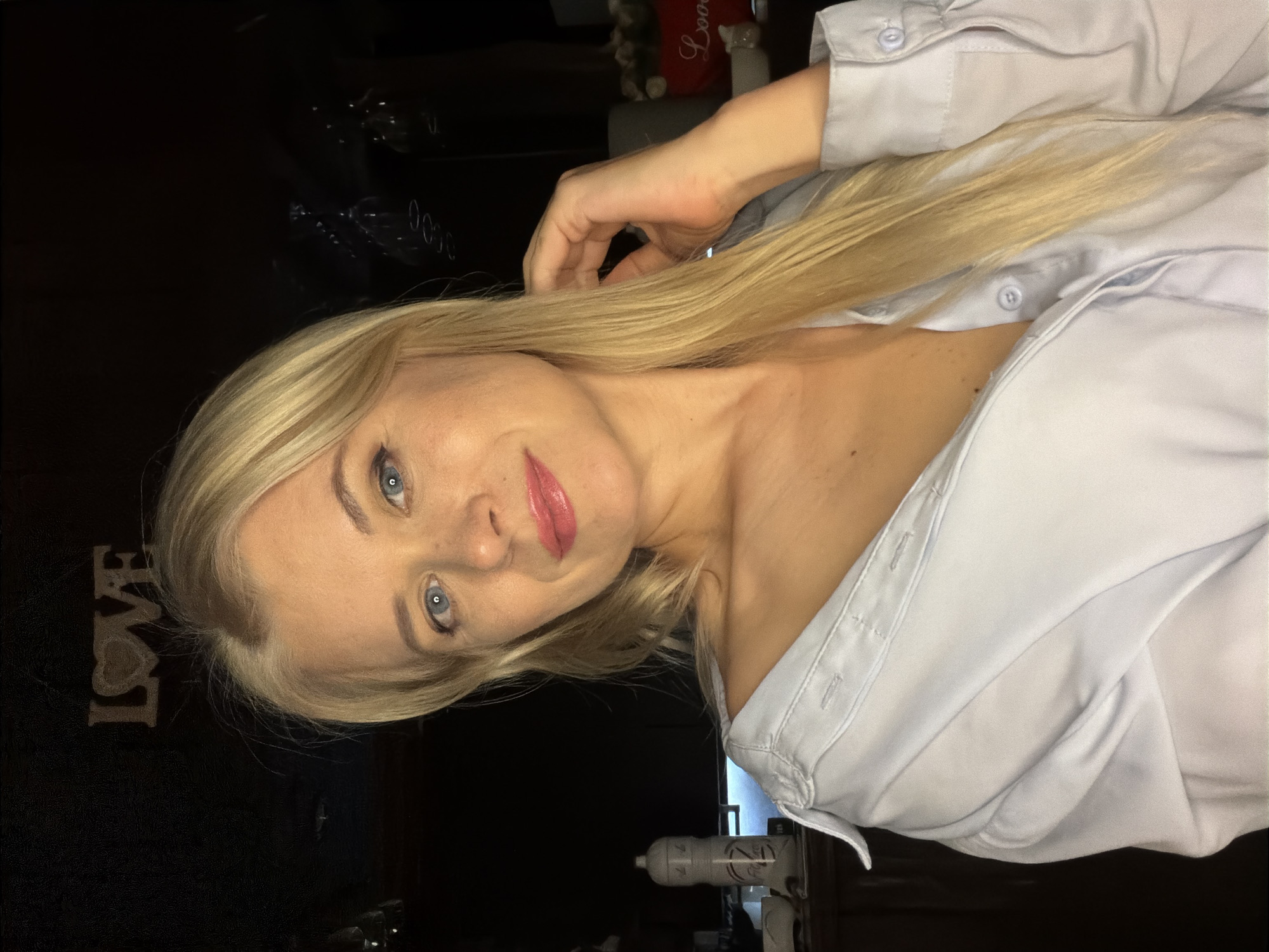 A woman with blonde hair and blue eyes poses for a photo, her head tilted and her gaze directed towards the camera. She is wearing an unbuttoned light blue collared shirt, revealing her décolletage. Her hair is long and straight, falling over her shoulder and partially obscuring her chest. She has on pink lipstick and her eyes are accentuated with eyeliner. Her right hand is raised, with her fingers gently touching her hair.
The background is dark, with the exception of some blurry elements and a sign with the word "LOVE" in capital letters, with a heart shape in place of the 'o'. The lighting appears to be artificial, possibly from indoor lights or a flash, creating a somewhat dramatic effect.
The image appears to be a selfie, taken from a slightly low angle, emphasizing the woman's face and upper body. There are no clear indications of the time of day or weather. The location context provided is Sochi, Russia, but there are no specific visual cues in the image to confirm this. The overall mood conveyed by the image is one of playful confidence or sensuality.