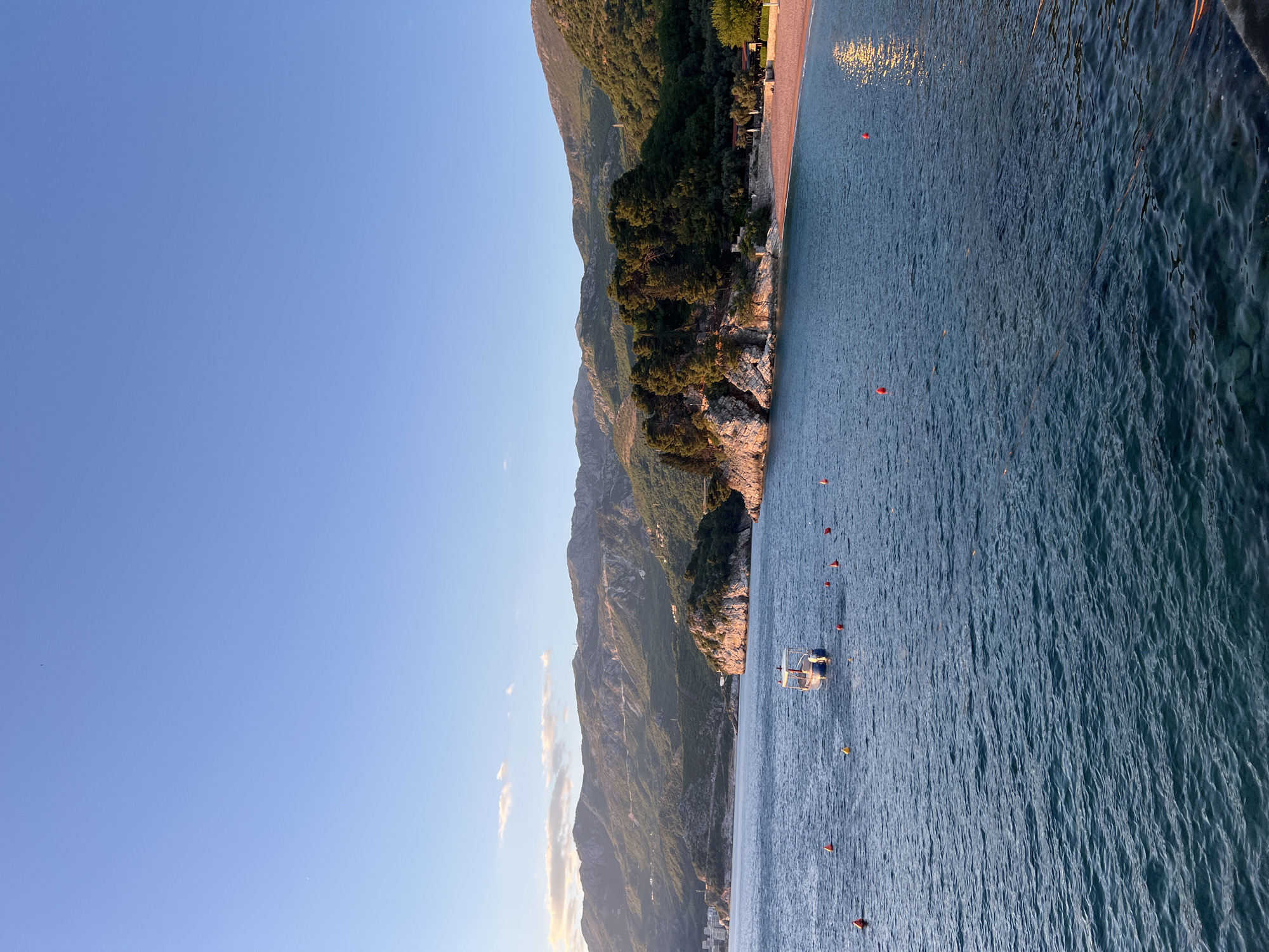 This image captures a serene coastal landscape in Sveti Stefan, Montenegro, during the late afternoon or golden hour. The scene is dominated by a majestic mountain range on the left, its rugged slopes covered in verdant green vegetation and trees, descending dramatically towards the sea. The setting sun casts a warm, golden glow on the mountain peaks and parts of the rocky coastline.
To the right, the calm, blue-green sea stretches out, its surface gently rippled, reflecting the soft light. A narrow strip of a light-colored beach is visible along the base of the mountains, also illuminated by the low sun. In the water, several red buoys are scattered, marking an area, and a small, white boat with a distinctive frame (possibly a parasailing boat) is visible. The sky above is a clear, bright blue, gradually lightening towards the horizon, with only a few faint, wispy clouds present. No people are clearly visible.
The overall atmosphere is peaceful and picturesque, highlighting the natural beauty of Montenegro's Adriatic coast. There is no visible text in the image.