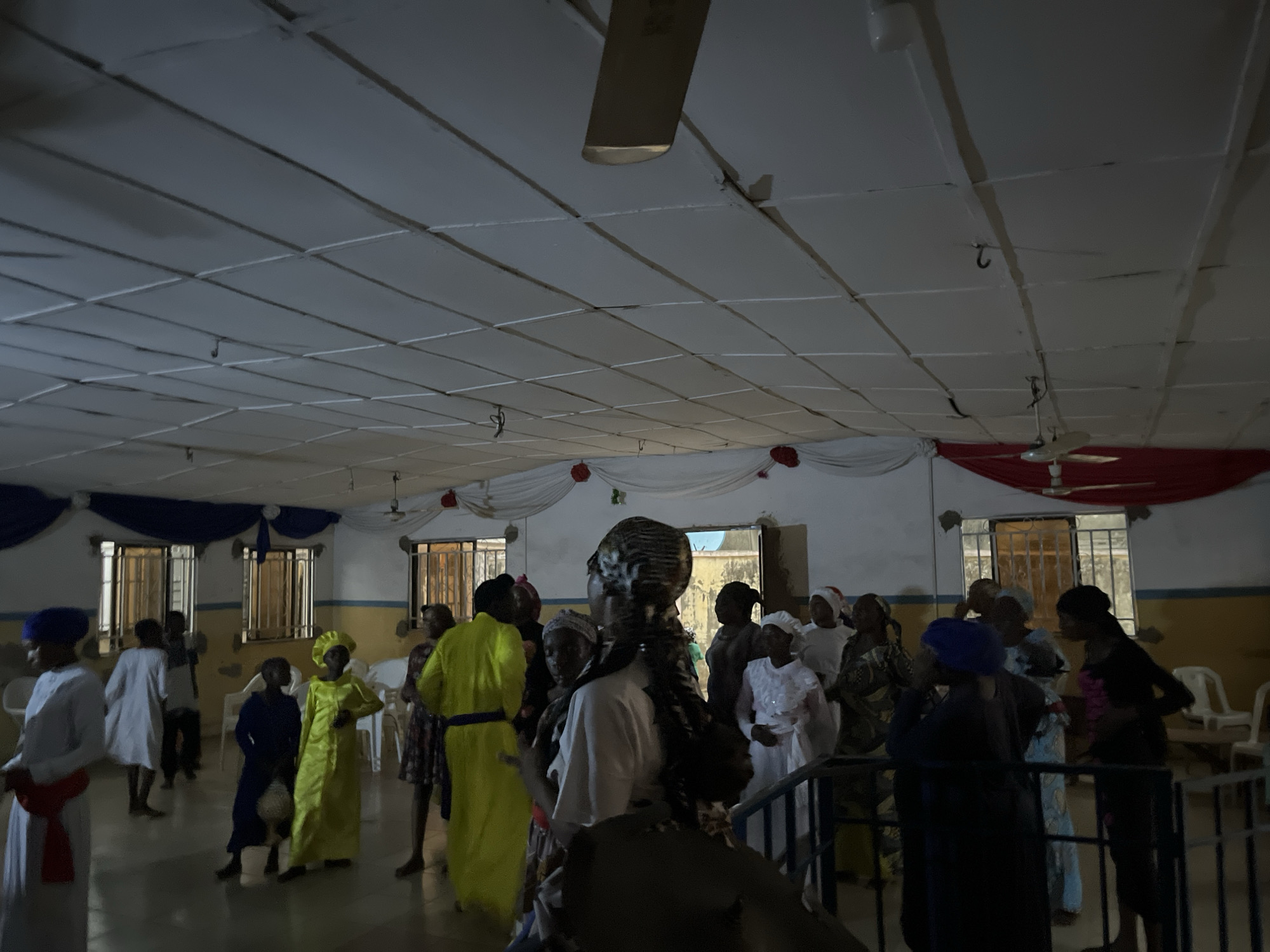 The image depicts an indoor gathering of approximately twenty to thirty individuals, predominantly women and children, within a large hall. The setting appears to be an event space, possibly a community or religious hall, indicated by the white acoustic tile ceiling with visible gridwork, overhead ceiling fans, and decorative fabric drapes in blue, white, and red along the upper walls. Red rosette accents are also present on the white drapery.
The walls are painted in two tones: white on the upper section and a light yellow or beige on the lower section, showing signs of wear and stains. Multiple rectangular windows with dark metal security grilles are visible on the far wall, providing a glimpse of a faint exterior. The floor is dark and somewhat reflective.
The individuals are dressed in various forms of attire, with many wearing long robes or dresses. Noteworthy clothing includes: a person in the left foreground wearing a white robe with a red sash and blue head covering; a child in the mid-left wearing a bright yellow robe and matching head covering; another child nearby in a dark blue robe holding a light-colored, spherical object. Several other individuals are dressed in white robes and head coverings, particularly in the mid-right. People are standing, walking, and engaging in low-level interaction, suggesting a social or communal event. White plastic chairs are visible stacked or arranged in the background. A dark metal railing is present in the right foreground, suggesting a raised platform or stairway. The overall lighting is dim, casting soft shadows across the scene. The location is an indoor event space within the city of Unknown, Unknown.