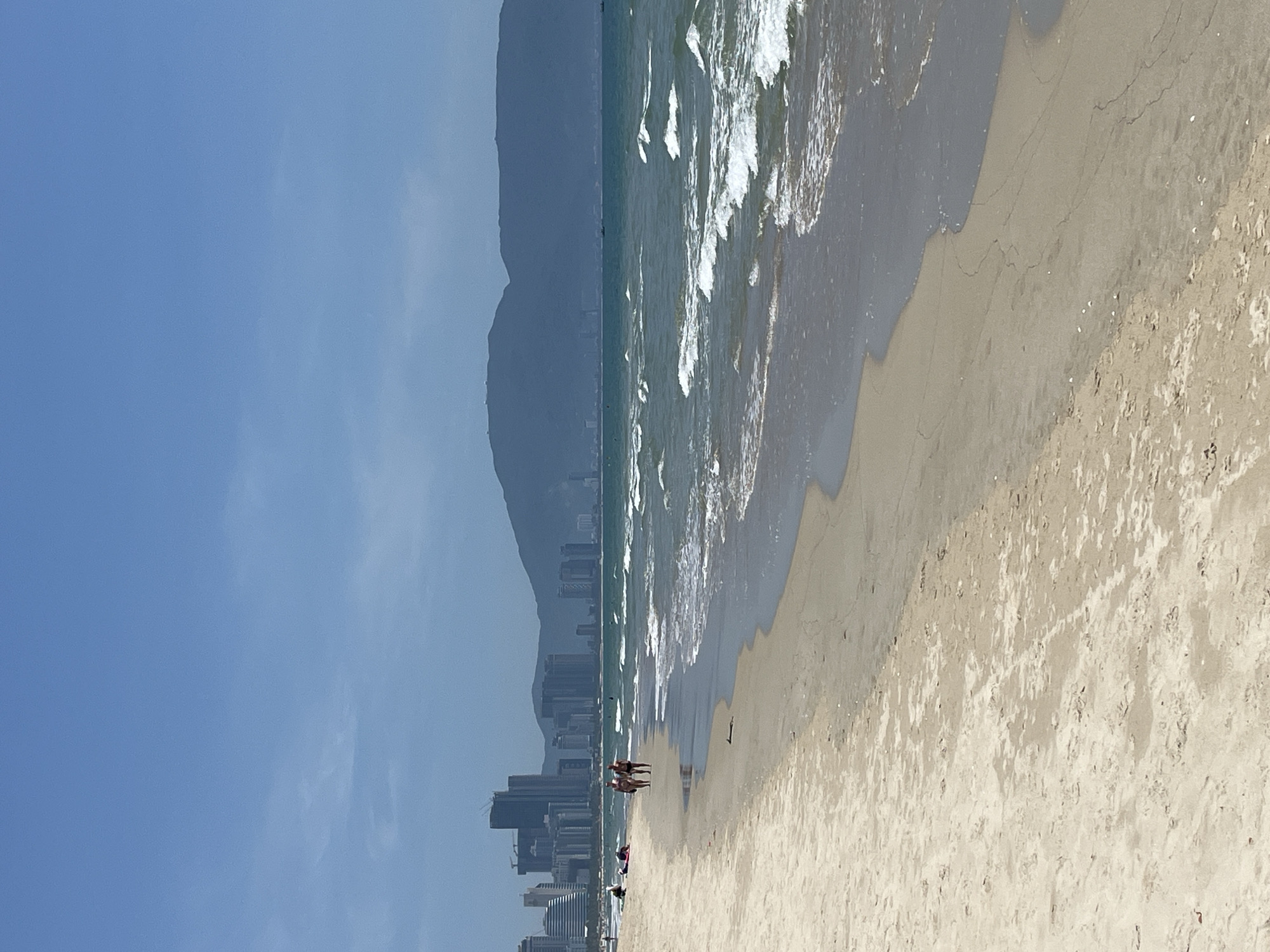 The image shows a beach in Đà Nẵng, Vietnam. The sky is blue with minimal cloud cover. In the distance, a mountain range is visible, obscured by haze. A line of buildings and skyscrapers marks the city skyline along the coastline. The ocean has waves breaking along the shoreline, with white foam contrasting against the green-blue water. The beach is sandy and light-colored. Several people are standing near the water's edge. The image is rotated 90 degrees counterclockwise.