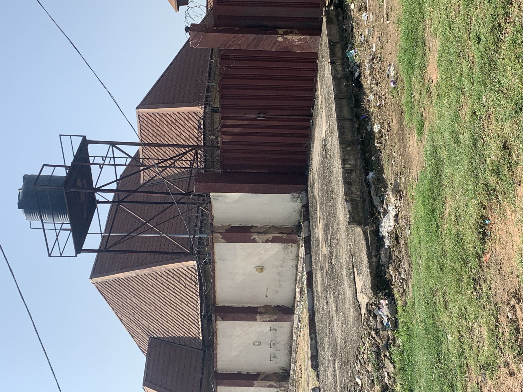 A low-angle, full outdoor shot captures a building's exterior with a dark green water tank elevated on a metal structure above a brown tiled roof. The roof is steeply pitched and appears to have several gables. A rusty metal gate, painted a dark reddish-brown, stands in front of a white concrete wall that has brown vertical columns and areas of peeling paint. Barbed wire is strung along the top of the wall and the gate. In the foreground, a cracked concrete path leads to the gate, with visible debris and trash scattered on the ground. The path is bordered by patchy green grass and bare, dry earth. Power lines stretch across the clear, pale blue sky.