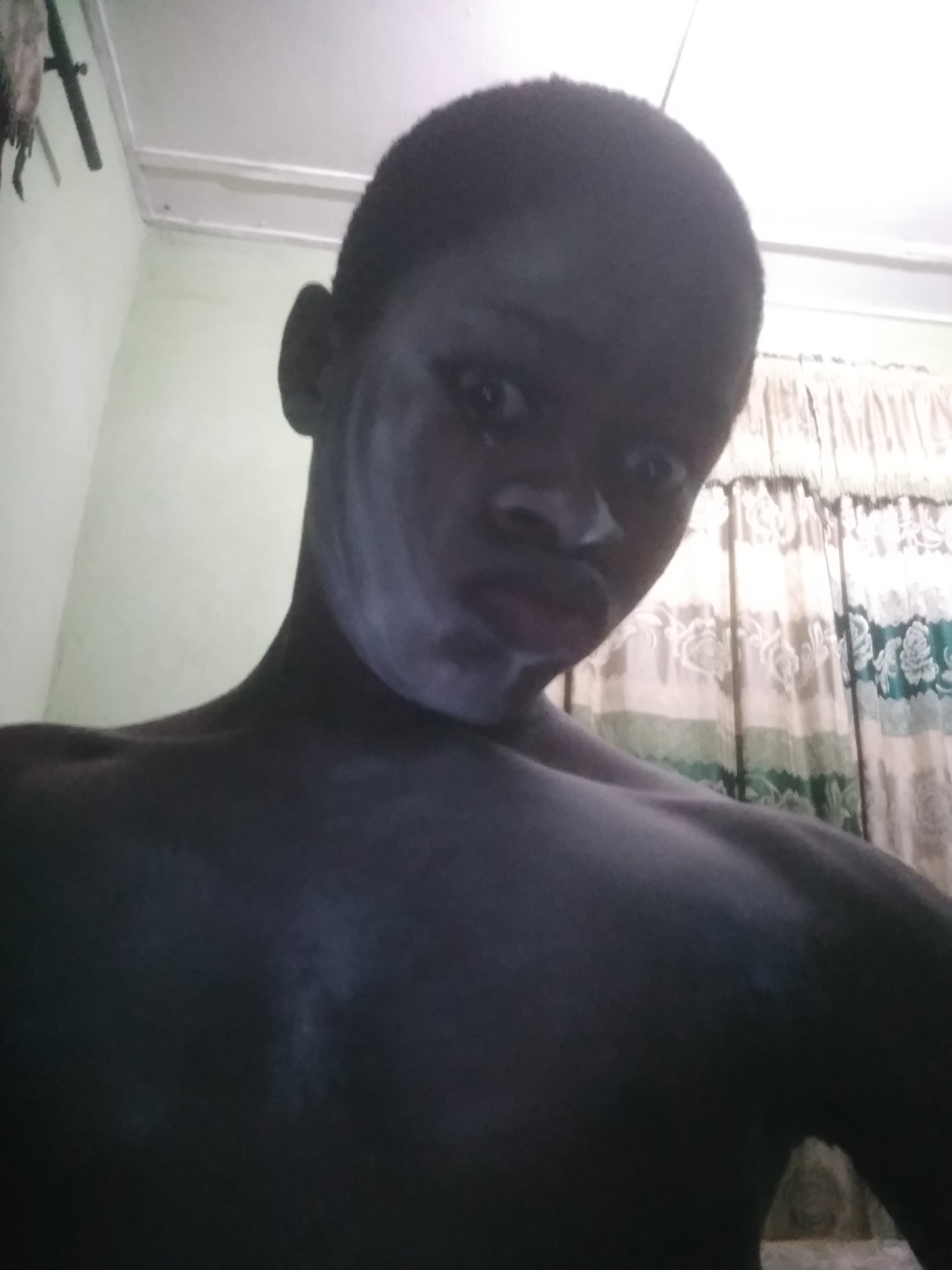 A dimly lit, close-up, low-angle self-portrait of a shirtless dark-skinned person with short hair. The person has a white substance on their face. The background consists of a pale green wall in the upper left, a white ceiling, and patterned curtains with green floral designs on the right. The exact location in Shangisha, Nigeria, cannot be determined from the provided image.