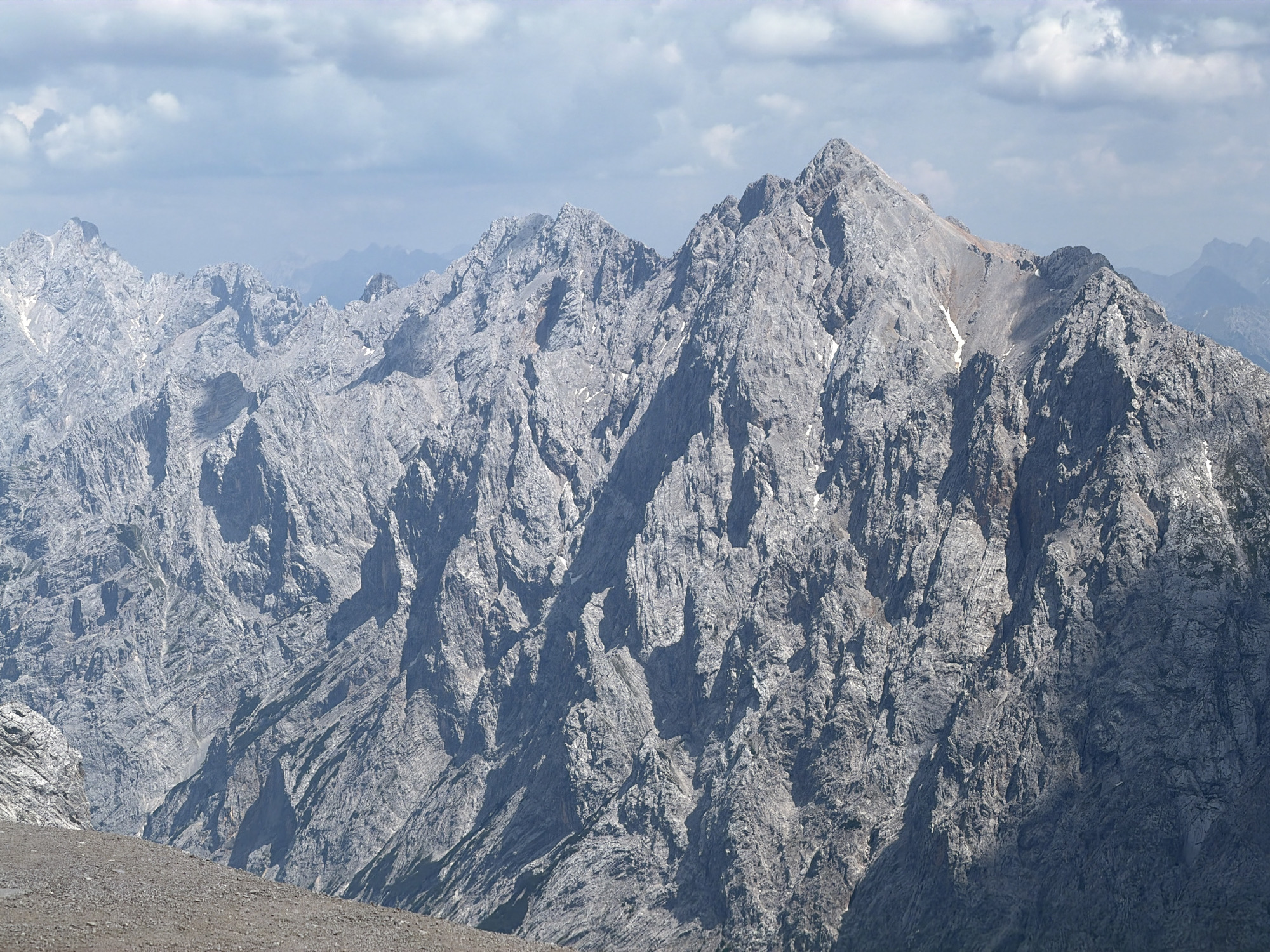 The image shows a rugged, mountainous landscape under a partly cloudy sky. The mountains are jagged and rocky, with steep slopes and sharp peaks. There are some patches of snow visible on the higher elevations. In the foreground, there is a gravelly, sloping area. The overall impression is one of a remote and wild natural environment.
While the image depicts a mountainous region, it is not specifically identifiable as being within the city of Garmisch-Partenkirchen, Germany. Garmisch-Partenkirchen is located in the Bavarian Alps and is known for its mountain scenery, including the Zugspitze, Germany's highest peak. However, this particular image does not contain enough specific landmarks or details to confirm its exact location within or near that city.