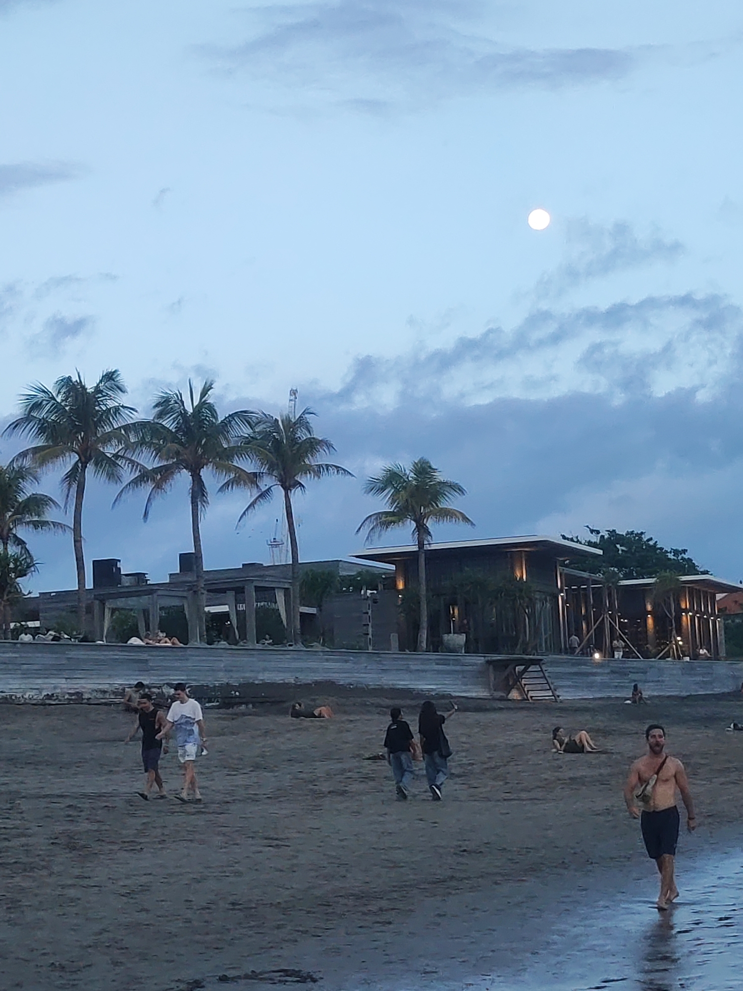 The image depicts a twilight scene at a beach in North Kuta, Indonesia. The sky is a pale blue-grey with scattered clouds, and a bright, full moon is visible in the upper right quadrant, indicating the early evening.
In the mid-ground, several tall palm trees stand prominently in front of and between modern, low-rise buildings. These structures, possibly a beach club or resort, feature contemporary architecture with flat roofs and subtle, warm lighting emanating from within and from outdoor fixtures. Some buildings incorporate wooden slats or pergolas, adding to their aesthetic.
The foreground is a sandy beach, which appears damp and reflective near the water's edge, a sliver of which is visible in the bottom right. Several people are present, engaged in various leisure activities. On the left, two men walk along the wet sand. Further to the right, two individuals walk away from the camera. A shirtless man wearing dark shorts and a fanny pack or sling bag walks towards the viewer on the right side of the frame, near the water. Other people are seen relaxing in the background, some lounging on the sand, and others near the lighted establishments.
The overall atmosphere is one of relaxed, casual leisure at a tropical beach destination as dusk settles. There is no visible text in the image.