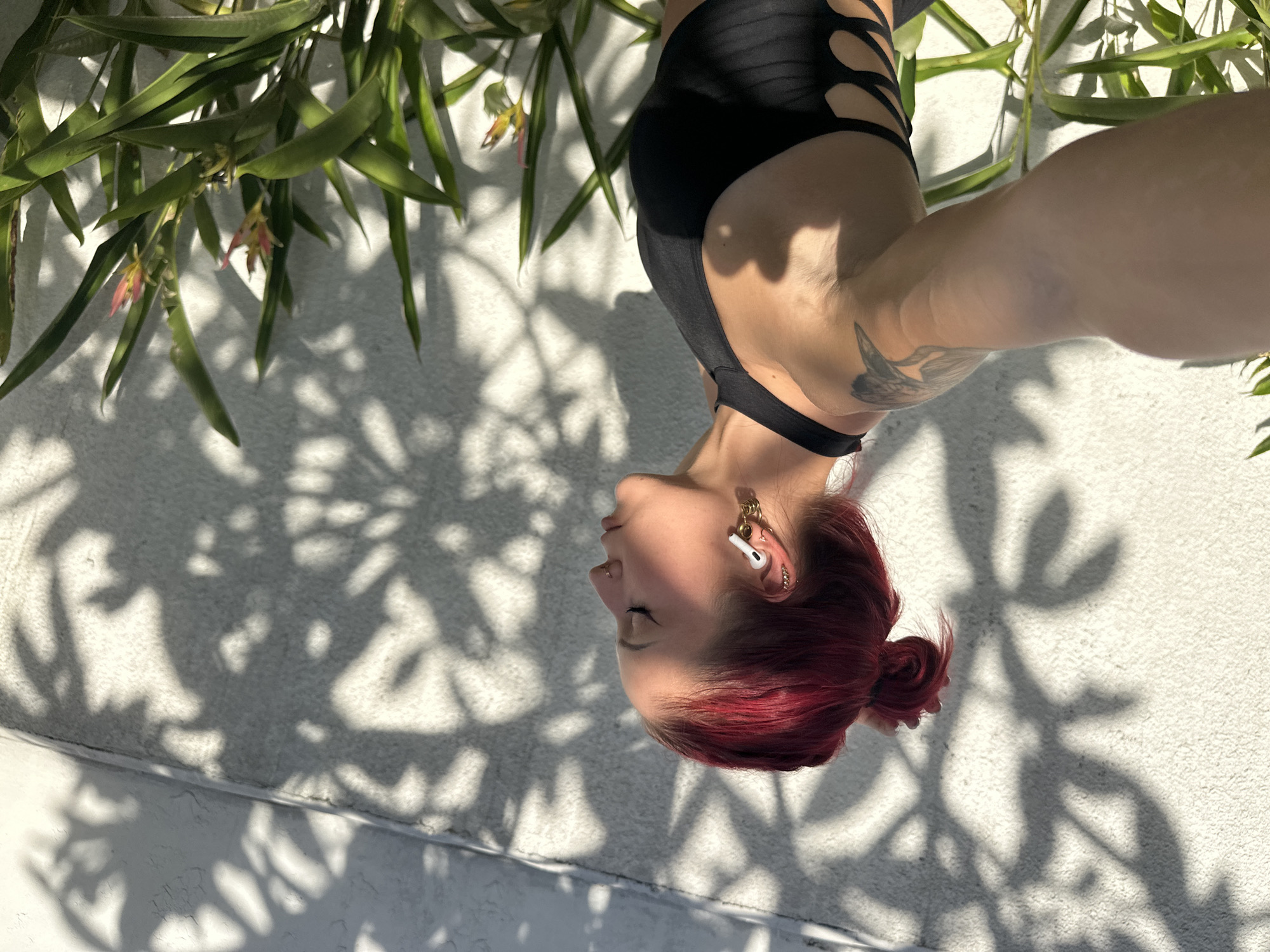 A medium-shot portrait captures a young woman positioned upside down, suggesting an acrobatic or suspended pose, set against a bright outdoor backdrop. The woman, with vibrant red hair styled in a bun, is seen in side profile with her eyes closed or cast downwards, conveying a sense of focus or tranquility. She wears a black top with cut-out details, white wireless earbuds, and multiple piercings on her ears and nose. A tattoo, possibly depicting a bird or wing, is visible on her right arm.
Above her head in the frame are lush green plants with small, delicate pink and yellow flowers. Behind and below her, a textured white wall serves as the main background. Strong, direct sunlight casts sharp, intricate shadows of the foliage across this wall, indicating a bright, sunny day. The scene takes place outdoors in North Kuta, Indonesia, with the interplay of light and shadow adding depth and visual interest to the composition. No visible text is present in the image.