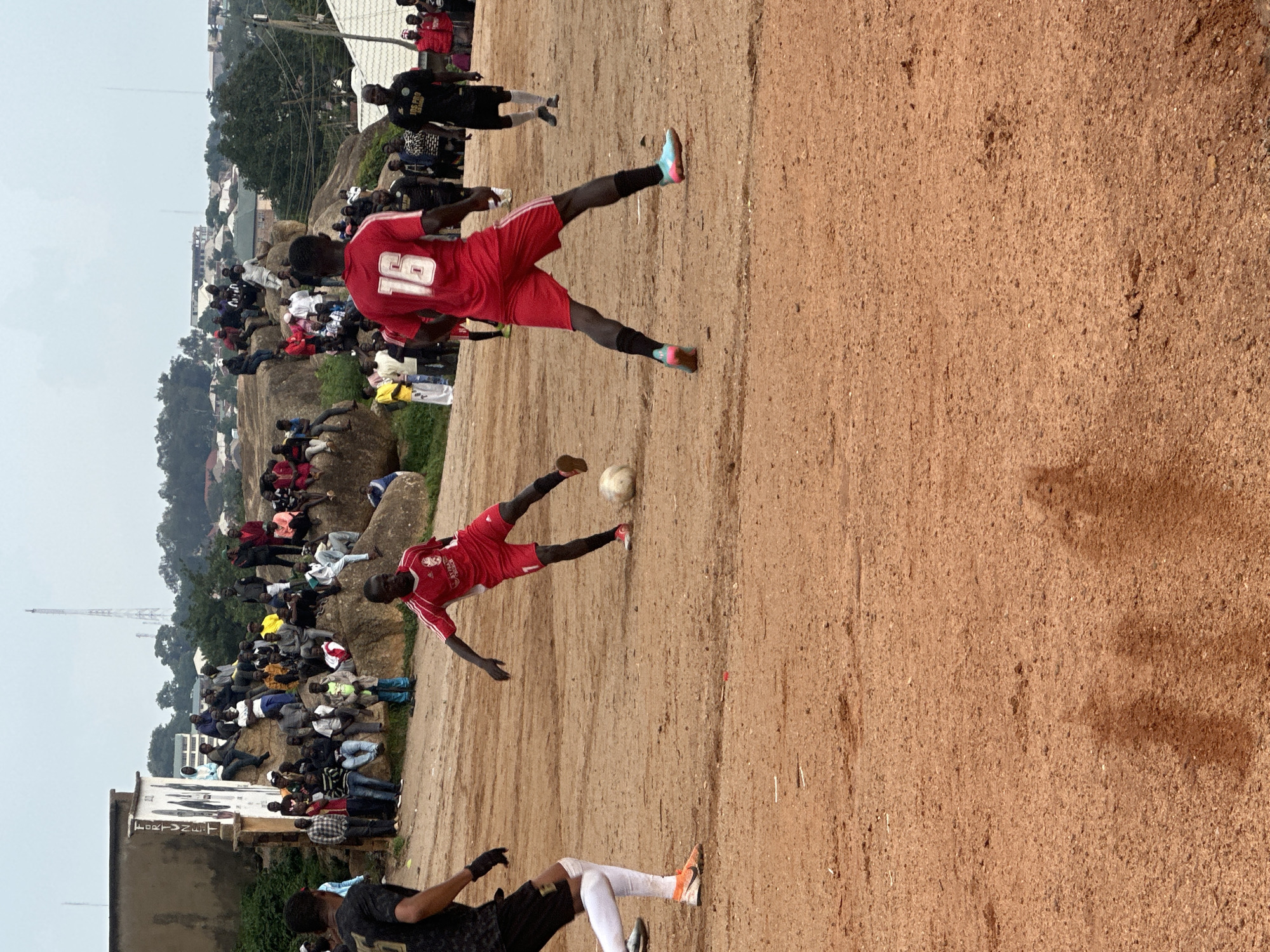 The image shows a group of people watching a soccer match on a dirt field in Jos, Nigeria. The spectators are seated on rocky outcrops and the ground surrounding the field. The players are wearing red and black uniforms. One player in a red uniform is kicking a soccer ball, while another player in a black uniform is positioned to receive it. The scene appears to be outdoors, with a cloudy sky suggesting overcast weather. In the background, there are buildings and trees, indicating a rural or semi-urban setting. A cell tower is visible in the distance. The overall atmosphere seems to be one of casual engagement, with people gathered to enjoy the game. The inscription "FORTUNET" is visible on a building in the foreground. The time of day is likely afternoon, based on the lighting.