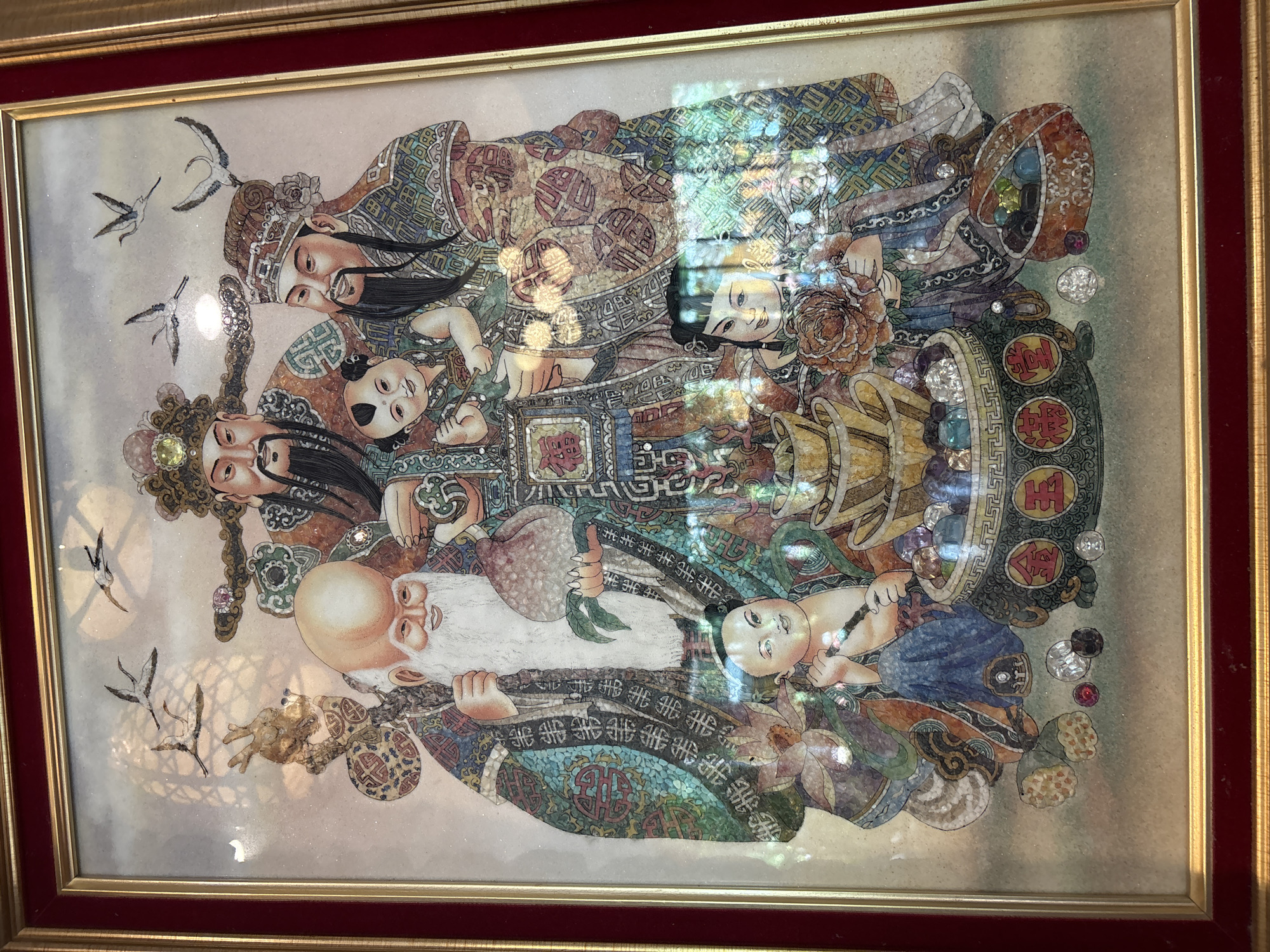 The image depicts a framed artwork, possibly a mosaic or shell inlay, featuring a traditional Chinese scene with multiple figures and symbolic objects. In the foreground, a large ornate pot or basin overflows with colorful gemstones and pearls, and is adorned with Chinese characters. To the left of this, a pile of lotus pods is visible. Two young children are prominent in the lower portion of the artwork; one sits and holds a decorative object, while another stands holding a bouquet of flowers. Behind them, three adult male figures are depicted. The central figure appears to be an elder with a long white beard, holding a peach. Two other figures flank him, one on each side, both wearing ornate headdresses and traditional robes. The background includes several flying cranes. The overall impression is one of prosperity and good fortune, common in Chinese iconography. The artwork is housed in a dark red frame with a gold inner border. Reflections from a light source are visible on the surface of the artwork, obscuring some details. There is no discernible indication of its location within the city of Chalong, Thailand; this artwork is presented as a standalone subject.