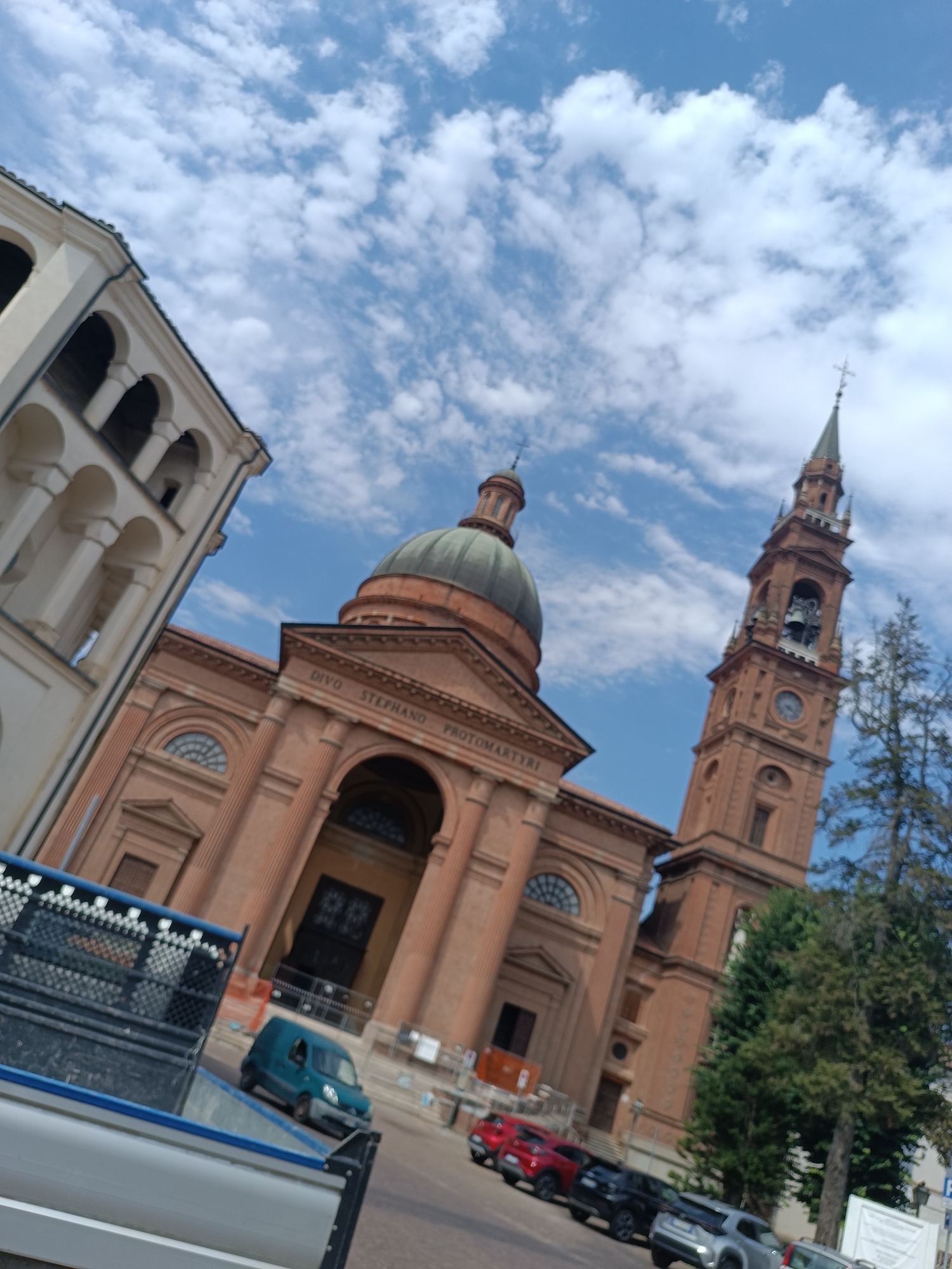 The image captures a view of the Duomo of Santa Maria Assunta in Casalmaggiore, Italy, on a partly cloudy day. The facade of the cathedral, constructed of brick, features a large dome and an adjacent bell tower with a clock. In the foreground, a white and blue truck bed is visible, and to its right, a teal van is parked on the street. Further down the street, a line of cars, including a red SUV, a black SUV, and a silver SUV, are parked. Lush green trees stand to the right of the bell tower, and a white sign is affixed to a pole in the lower right corner. The sky is a bright blue, dotted with white, fluffy clouds.