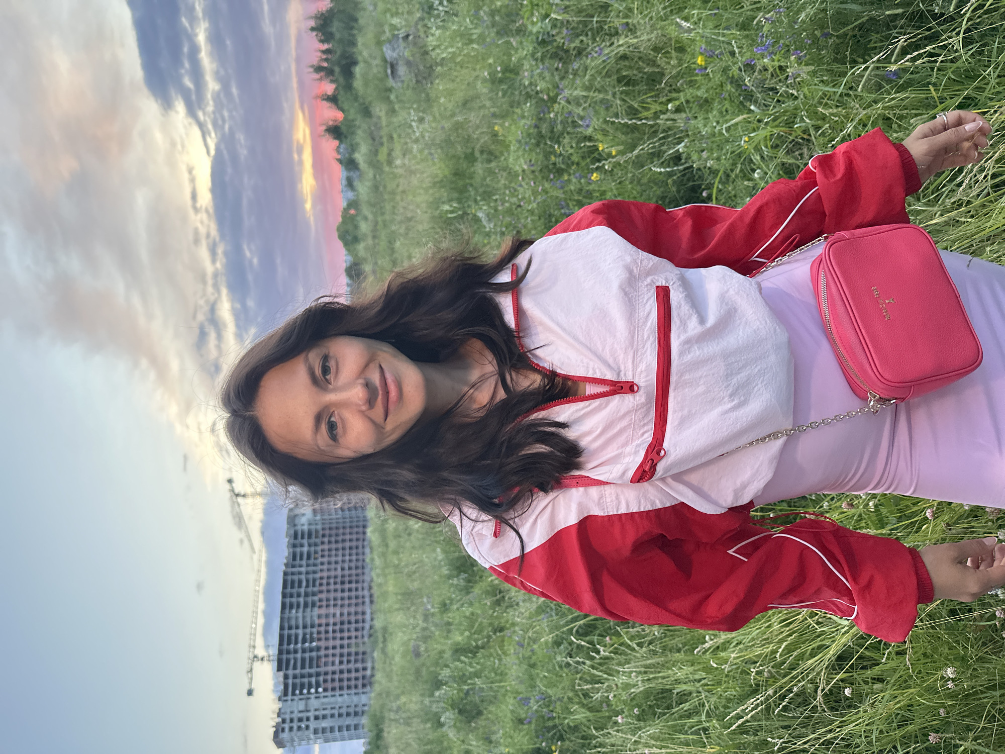 A young woman with long, dark, wavy hair stands in a grassy field at dusk. She is wearing a red and white quarter-zip jacket over a pink dress and a small pink crossbody bag. In the background, a large, unfinished building with cranes is visible, along with trees and a colorful sky. The location is identified as Murino, Russia, specifically near a construction site in a rural or undeveloped area on the outskirts of the city.